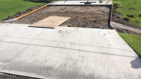 Concrete driveway with a section of dirt preparation.  A piece of plywood lies on the dirt.