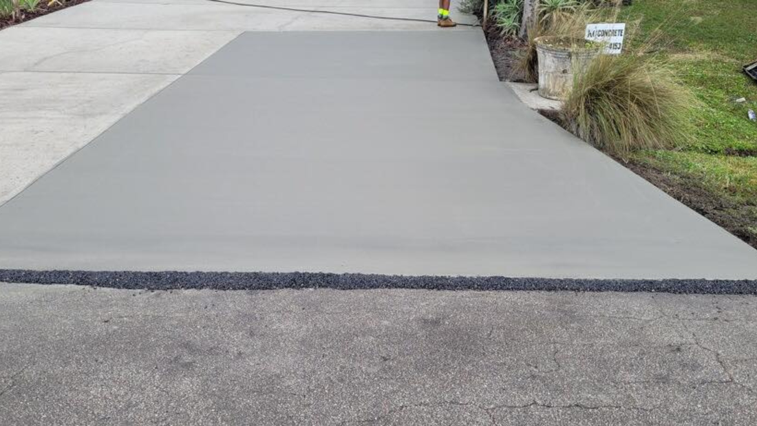 Newly poured concrete driveway extension, transitioning from asphalt, bordered by grass and landscaping.