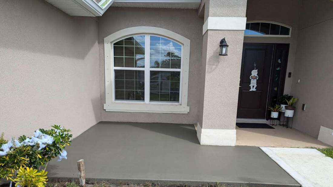 Front porch with freshly poured concrete, arched window, dark brown door, and greenery.