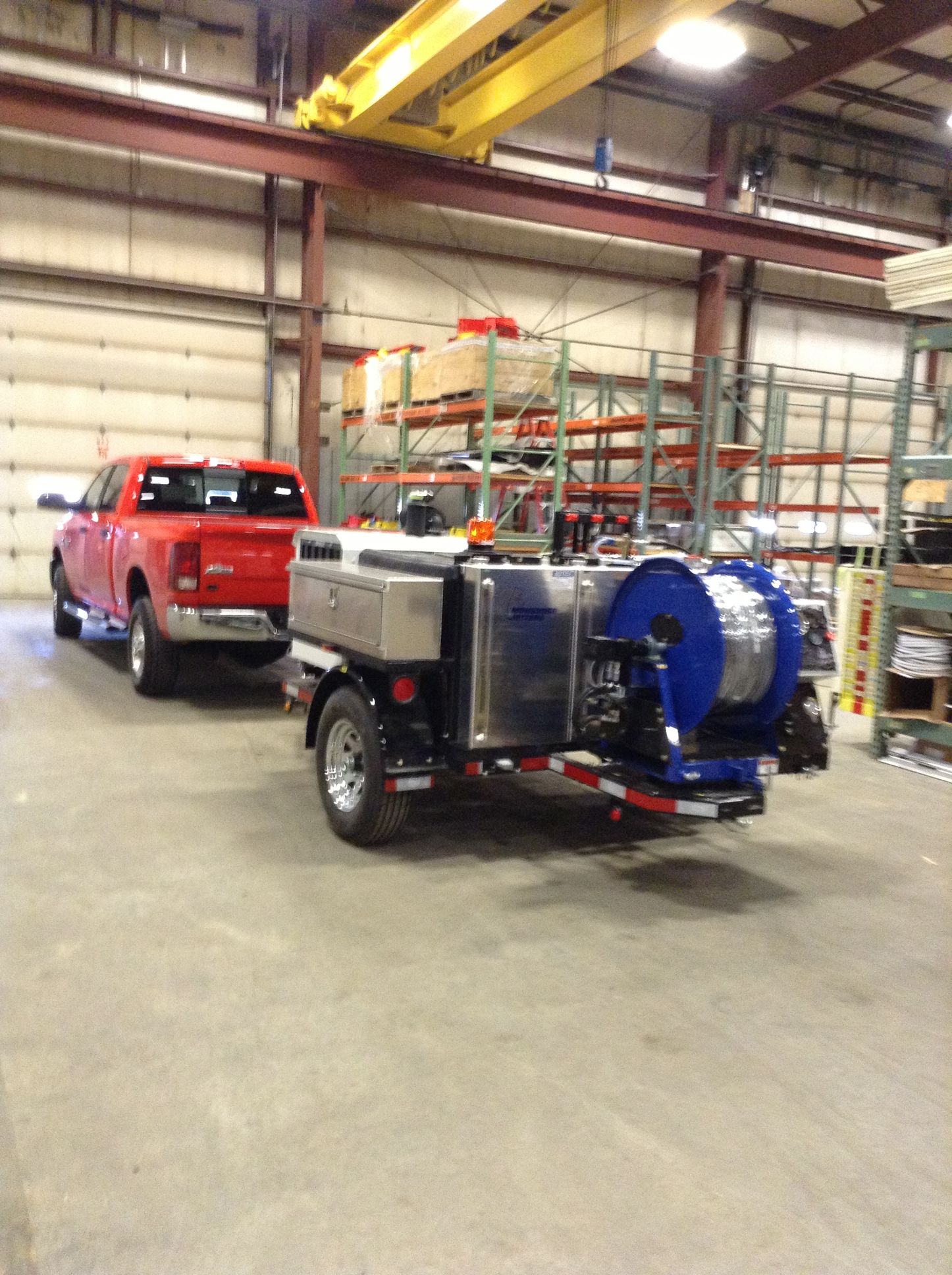 Red pickup truck towing a blue and silver trailer with a large blue hose reel inside a warehouse.