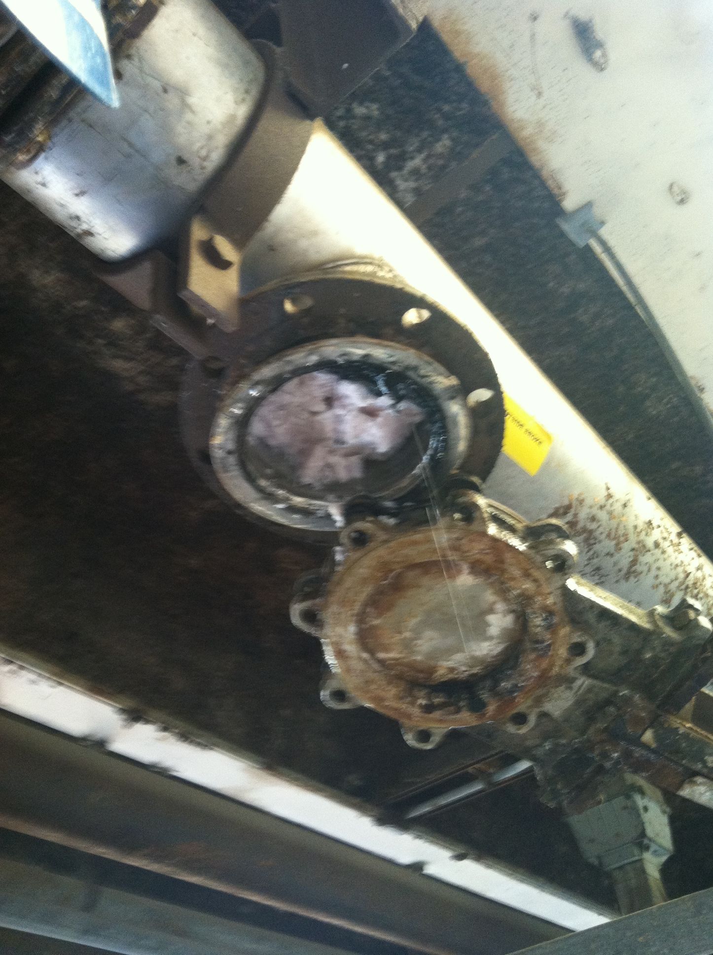 Corroded industrial valve connected to a metal cylinder, beneath a metal structure.