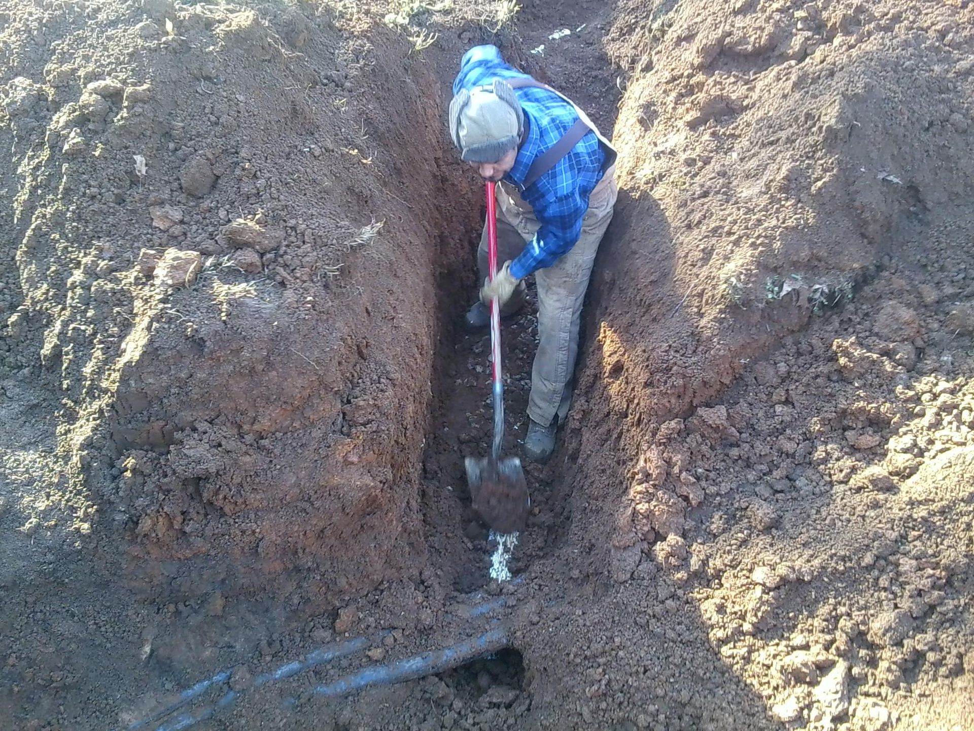 Person digging in a narrow trench with a shovel, surrounded by dirt.