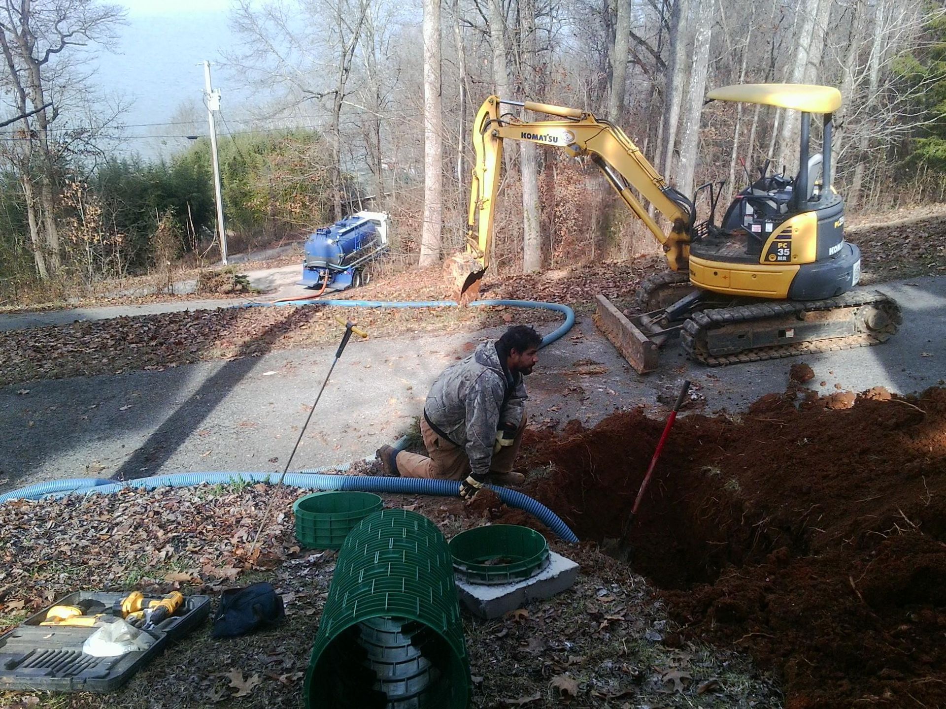 Man installing septic system with excavator. Earthwork in progress near trees and a blue tanker.