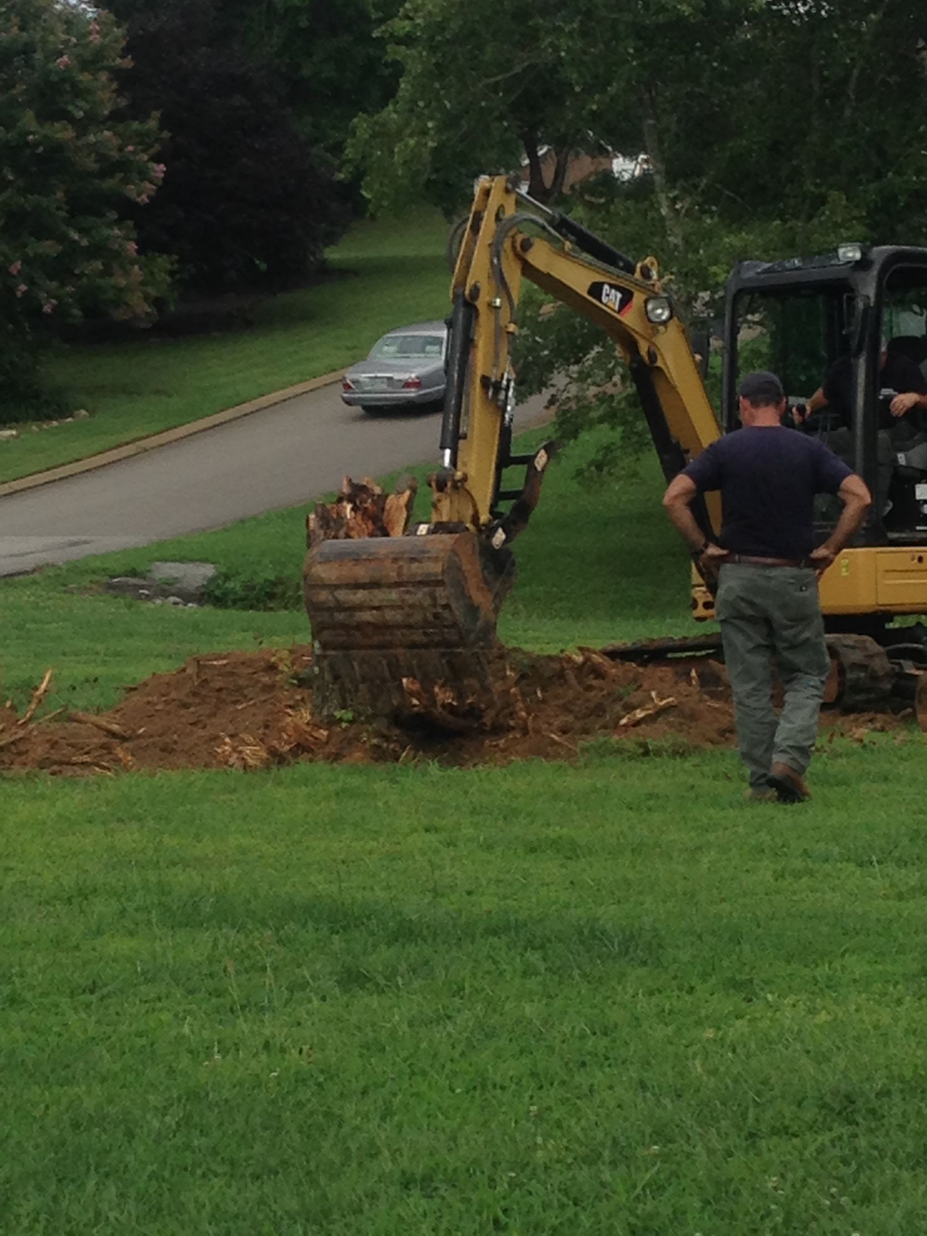 A man watches a yellow excavator dig in a yard near a road, dirt pile, and green grass.