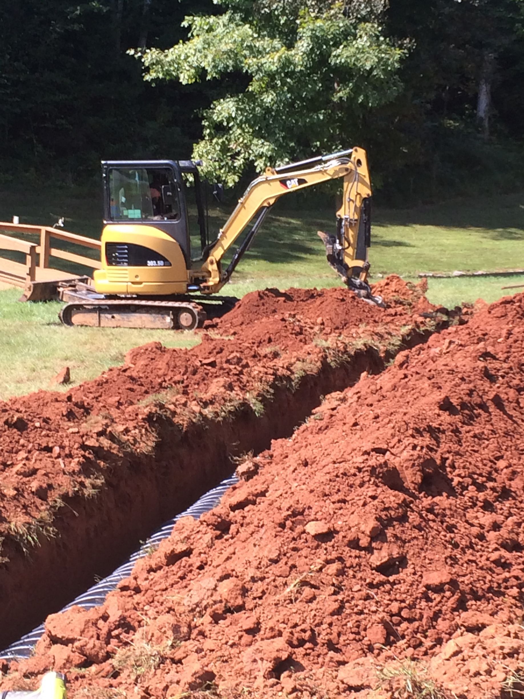 Yellow excavator digging a trench in red soil on a grassy property. A fence and trees are visible in the background.