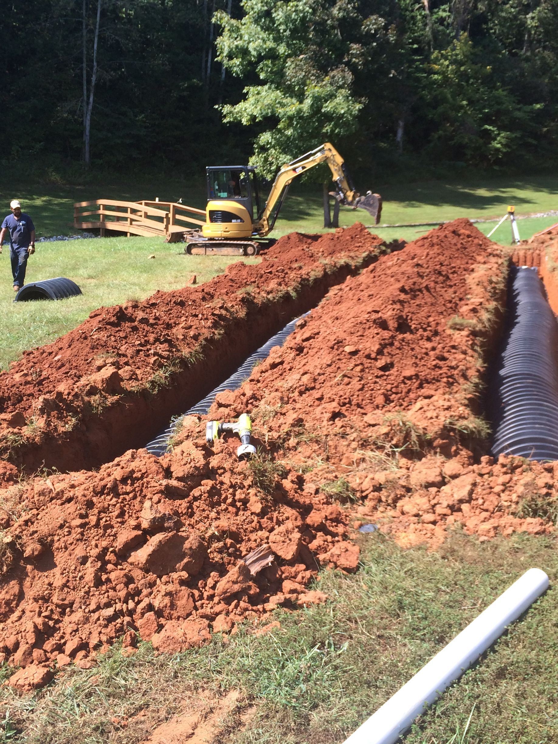 Excavator digging trenches in a grassy field; workers nearby, some using tools.