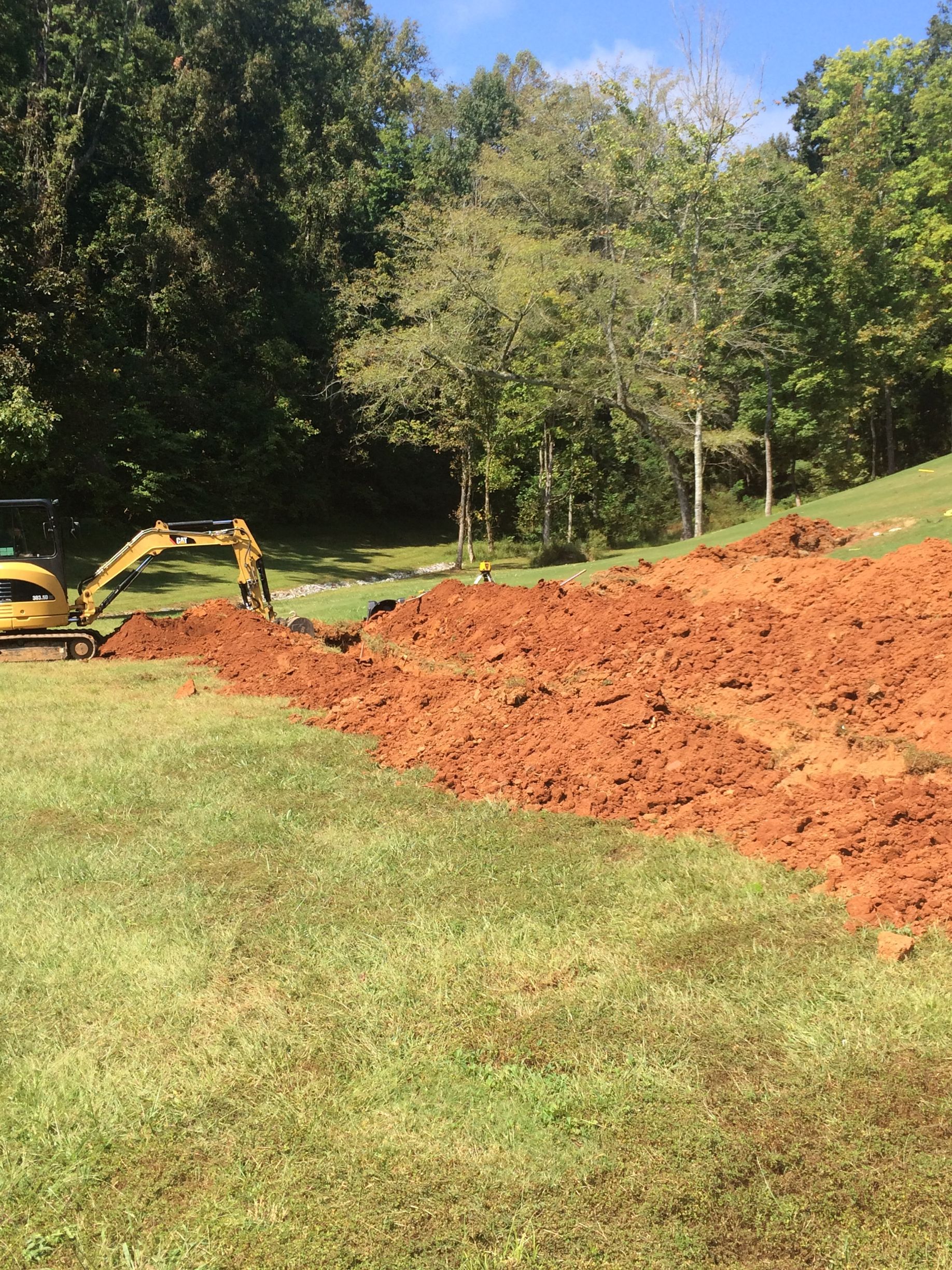 An excavator digs a trench in a grassy field with trees in the background. Brown soil is piled up.