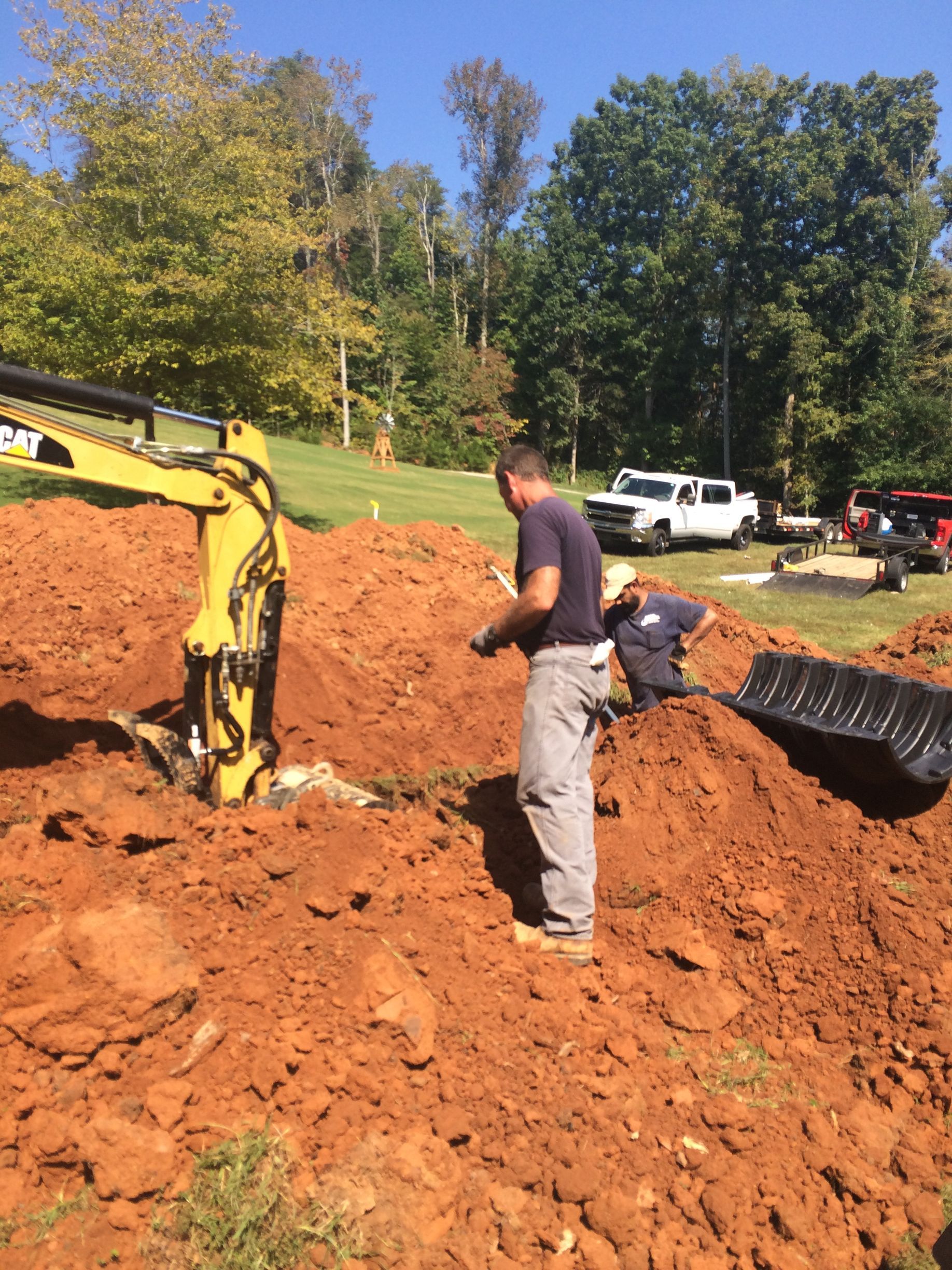 Two people working with an excavator and a black container on a dirt hillside. Sunny day.