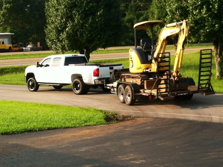 White truck towing a trailer with a yellow excavator on a paved road with green grass.