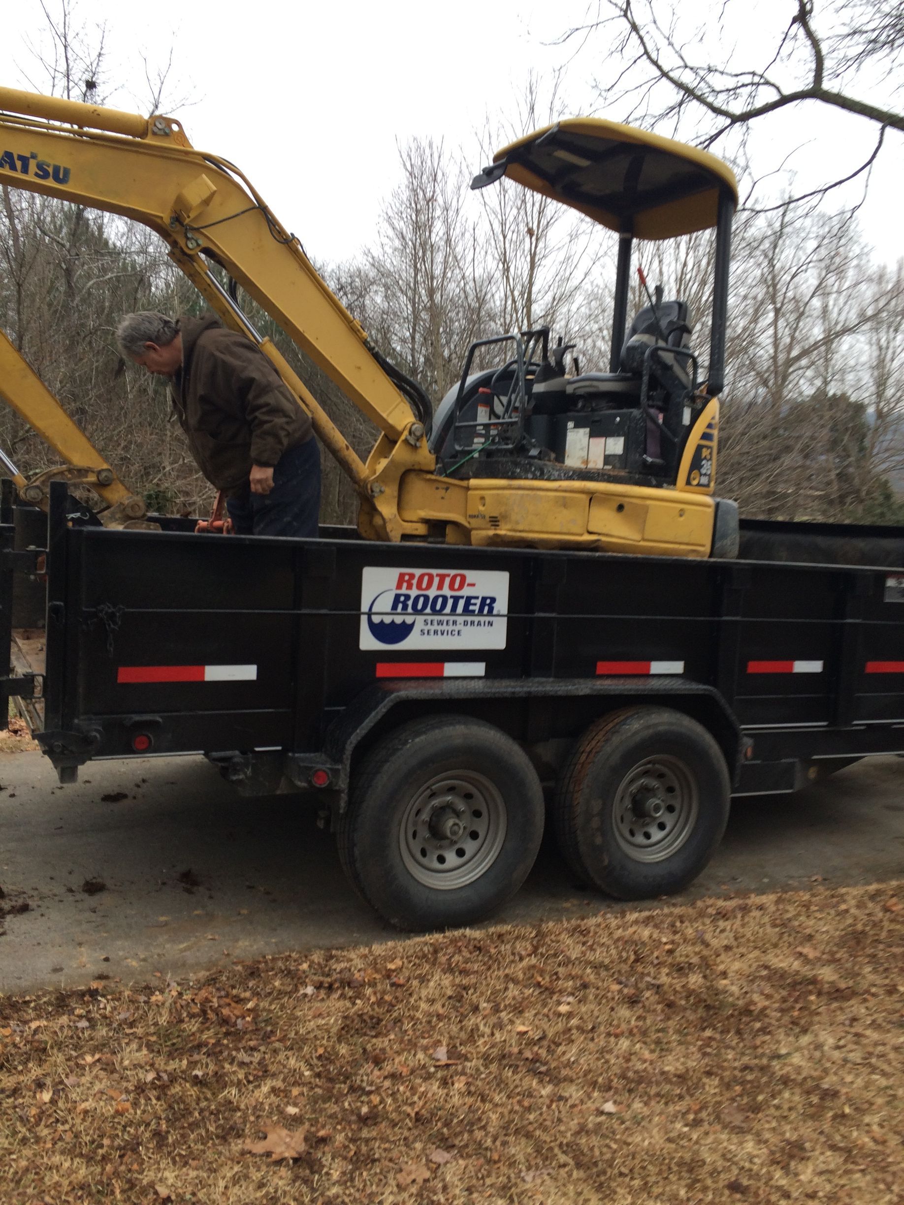 Person loading a yellow excavator onto a black trailer. The trailer has a logo. Overcast day.
