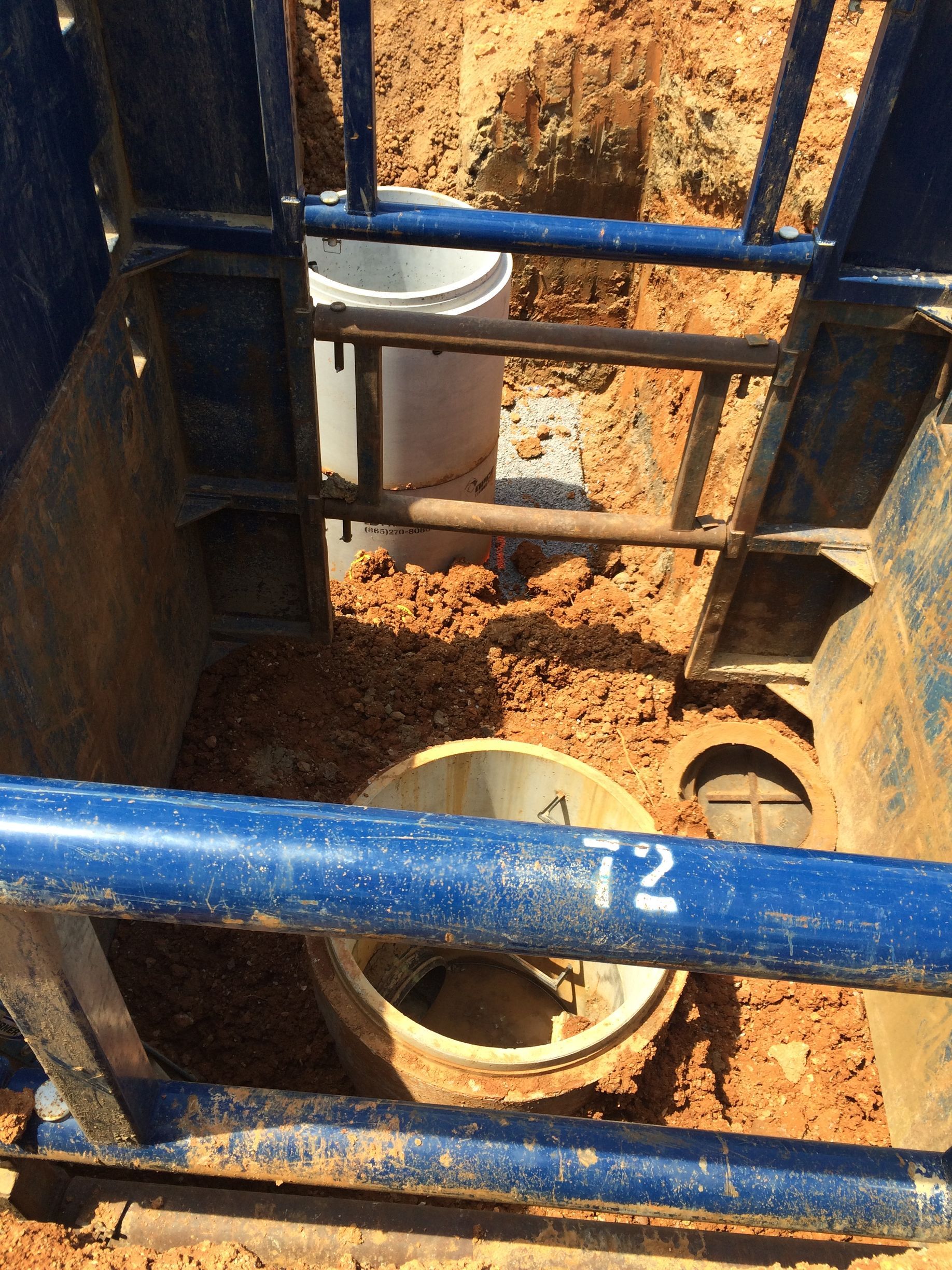 A trench with blue safety frame. Inside, a bucket and pipe are surrounded by dirt and gravel.