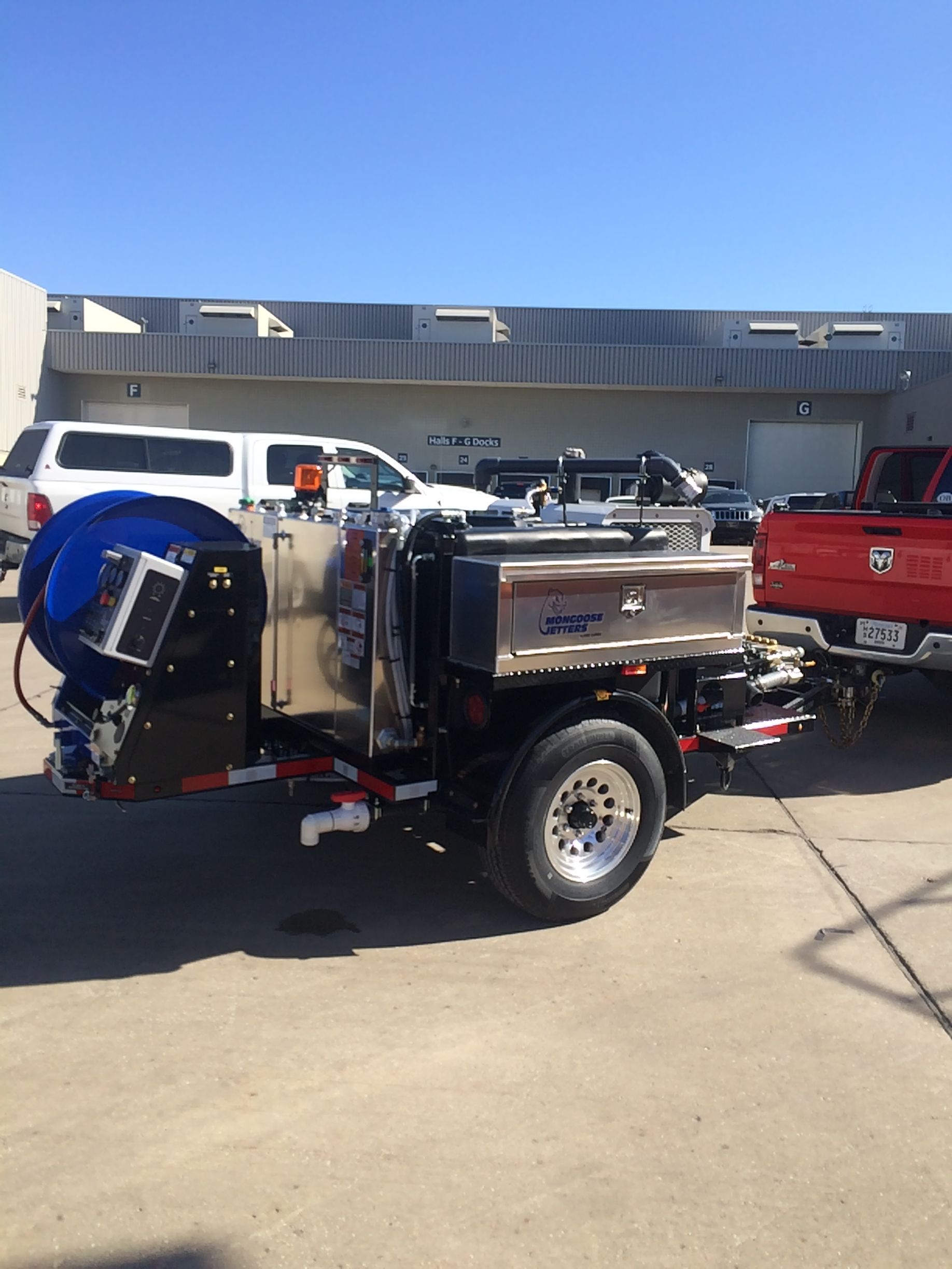 Truck-mounted jetting machine parked outside; blue hose reel, stainless steel tanks, and a red pickup truck are visible.