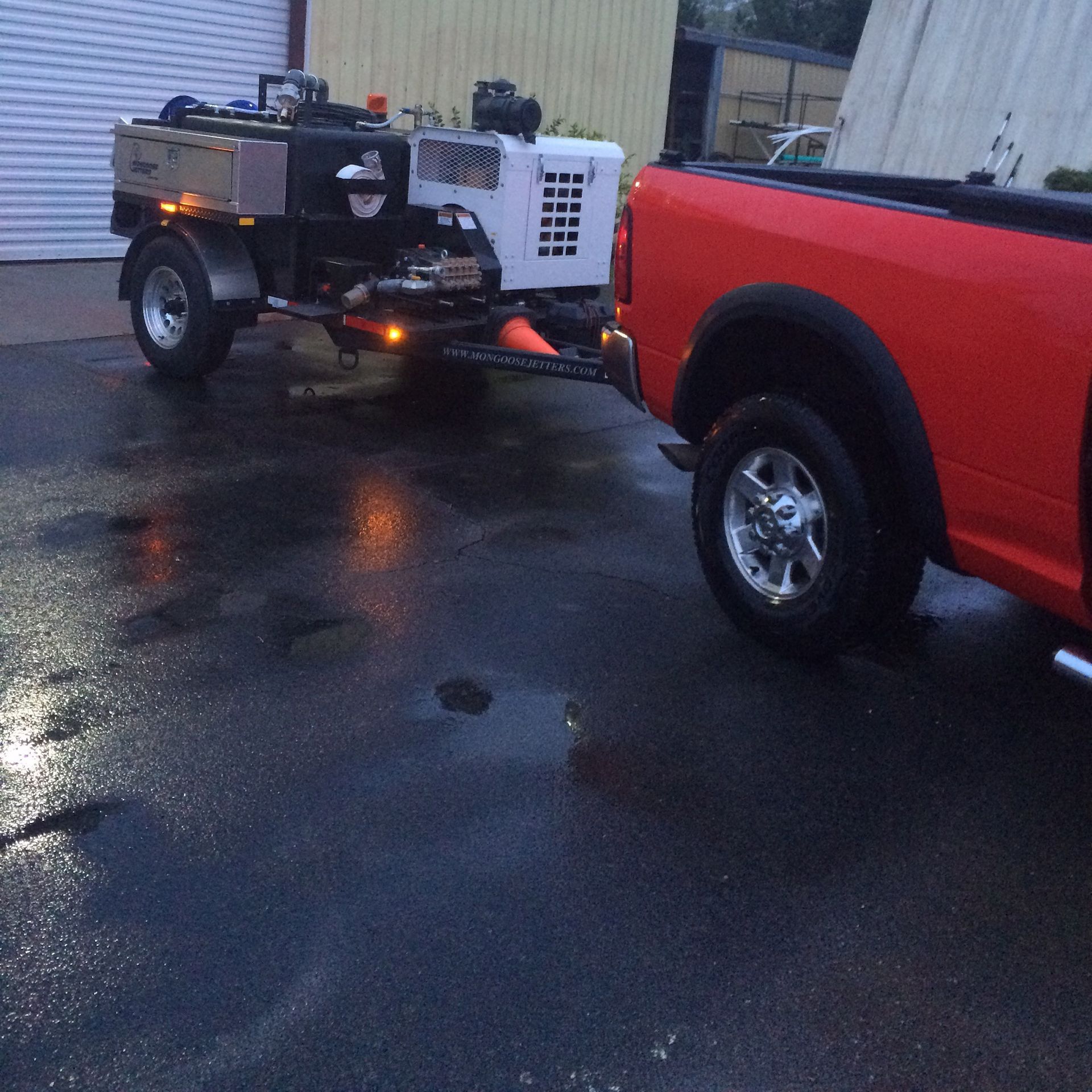 Red truck towing a trailer with a white machine; both parked on wet asphalt.