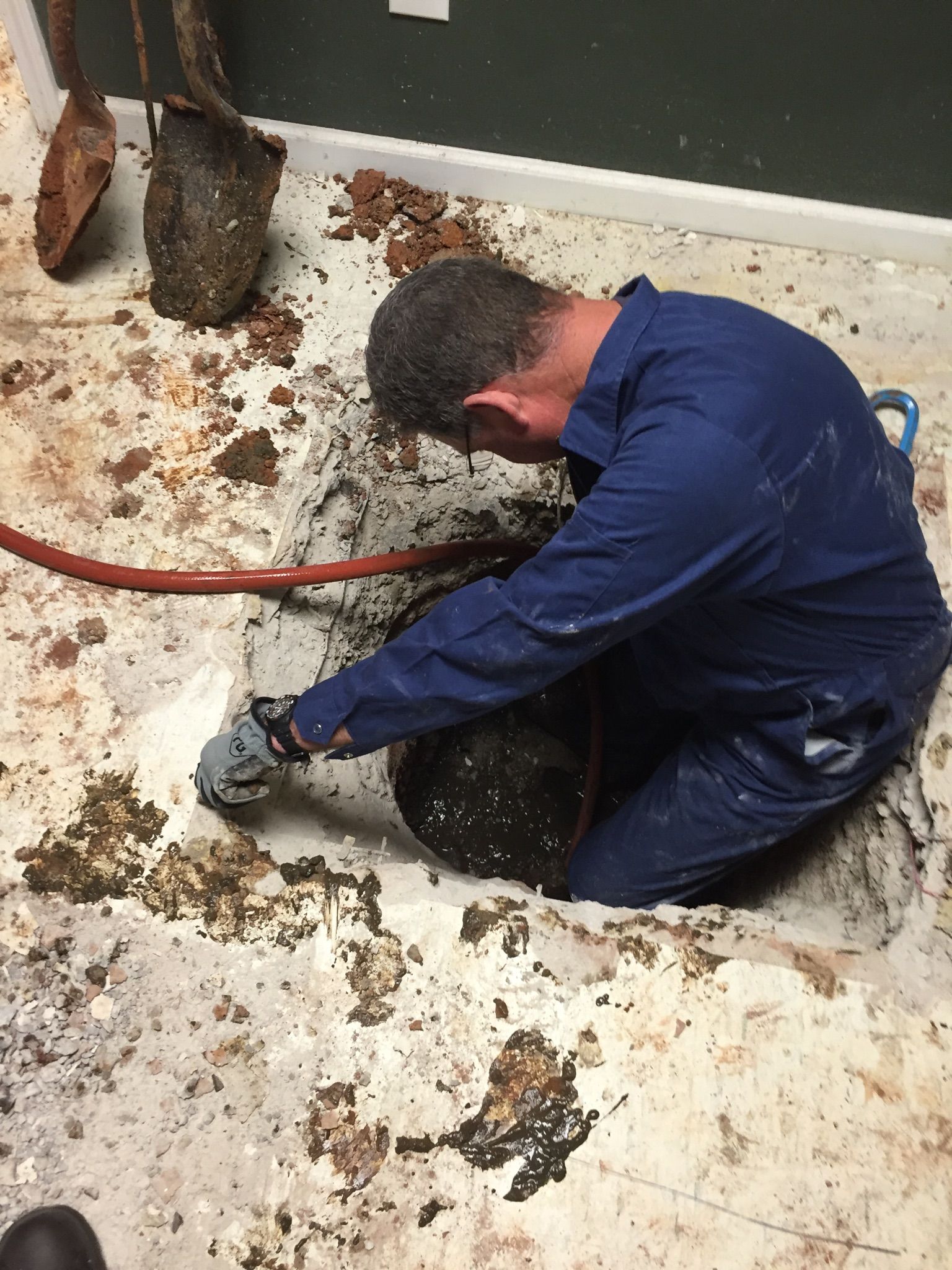 Man in blue jumpsuit works in a hole in a dirty floor, likely a repair. A hose and tools are present.