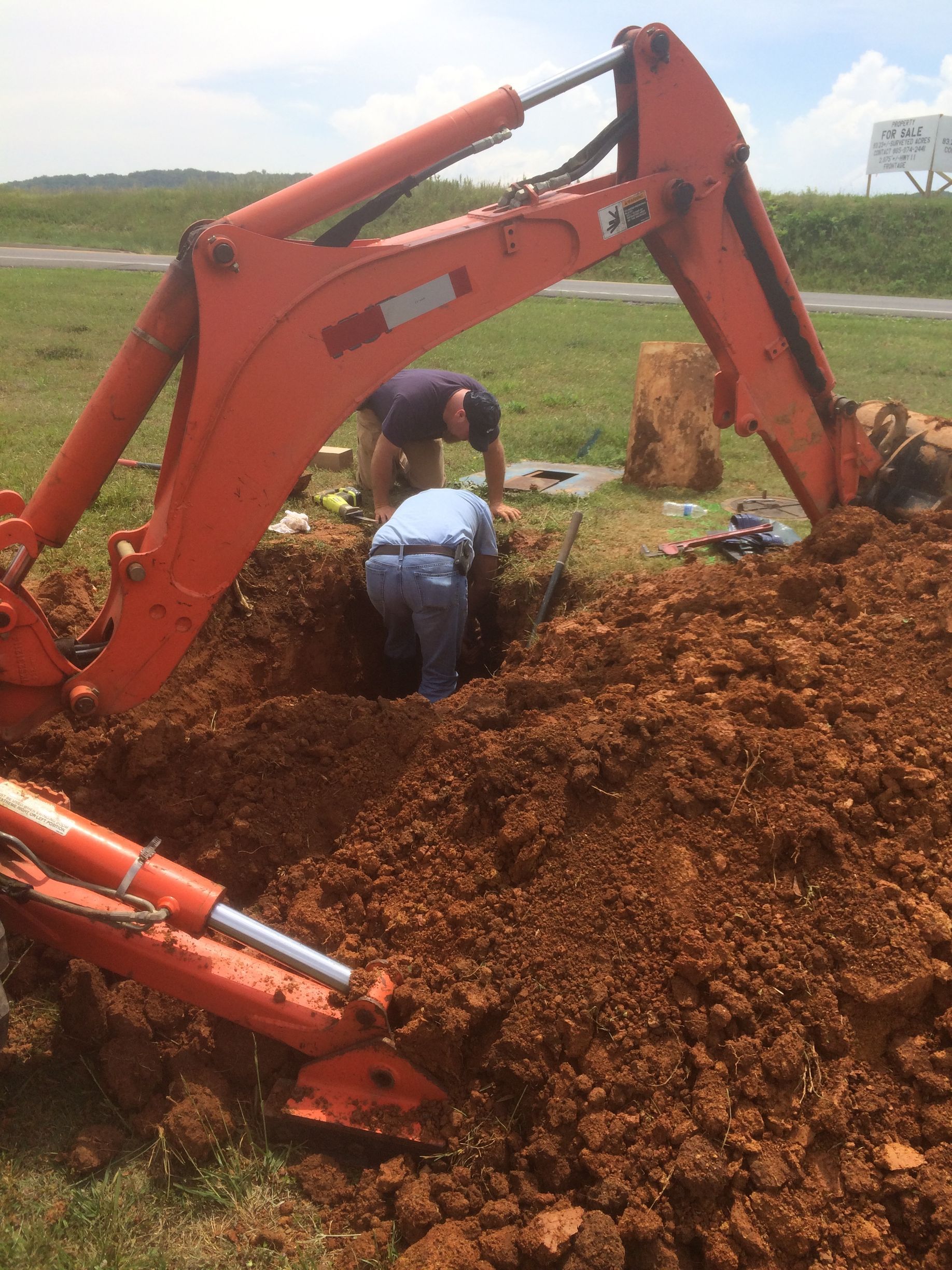 Orange excavator and two people digging in a hole in the ground outdoors.