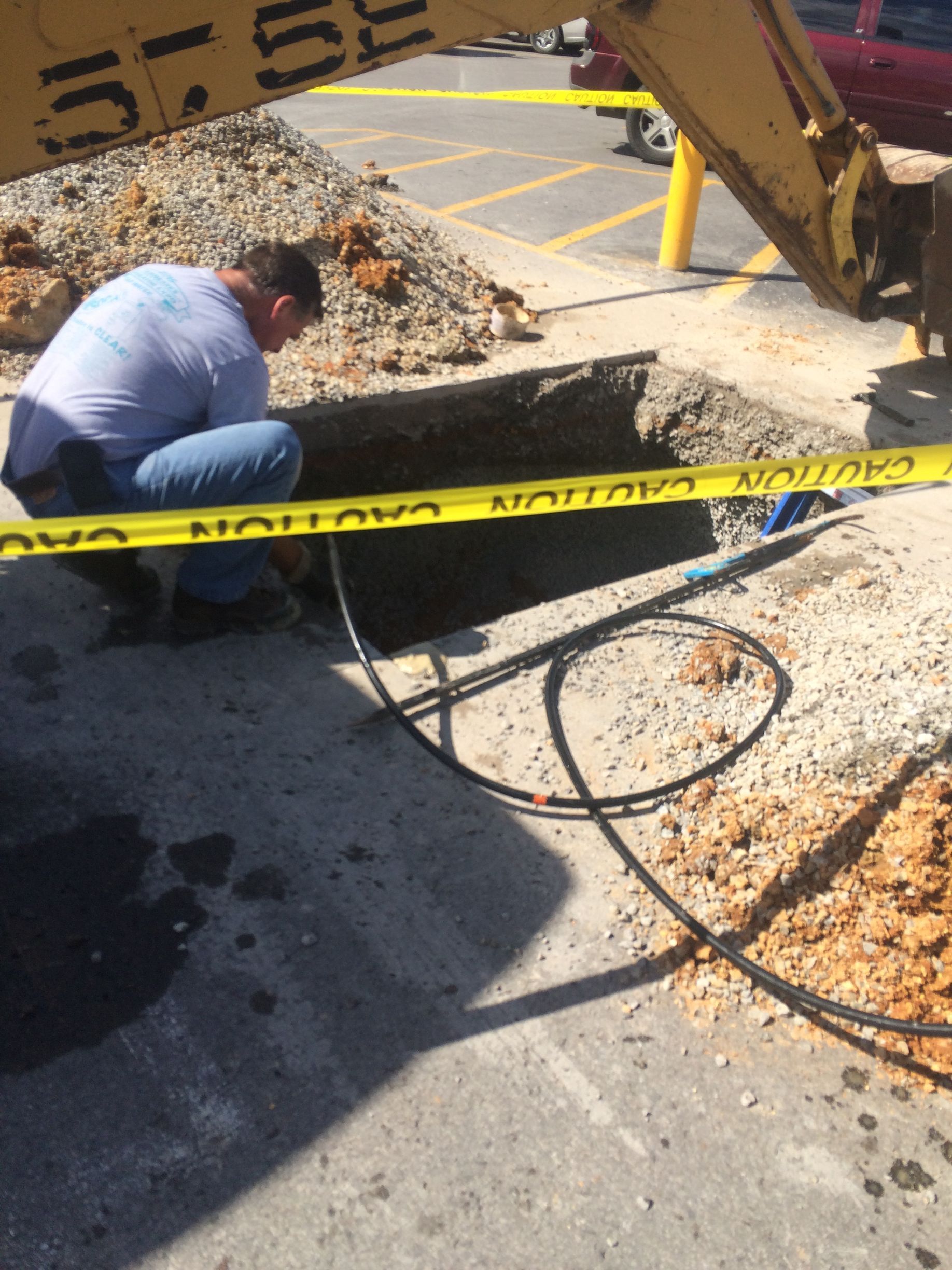 Man working near open trench with equipment and caution tape. Asphalt and dirt are visible.