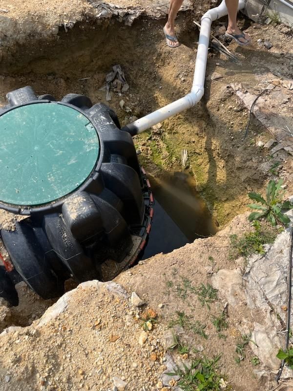 Black septic tank in a hole with a white pipe leading into it. Green lid is open.