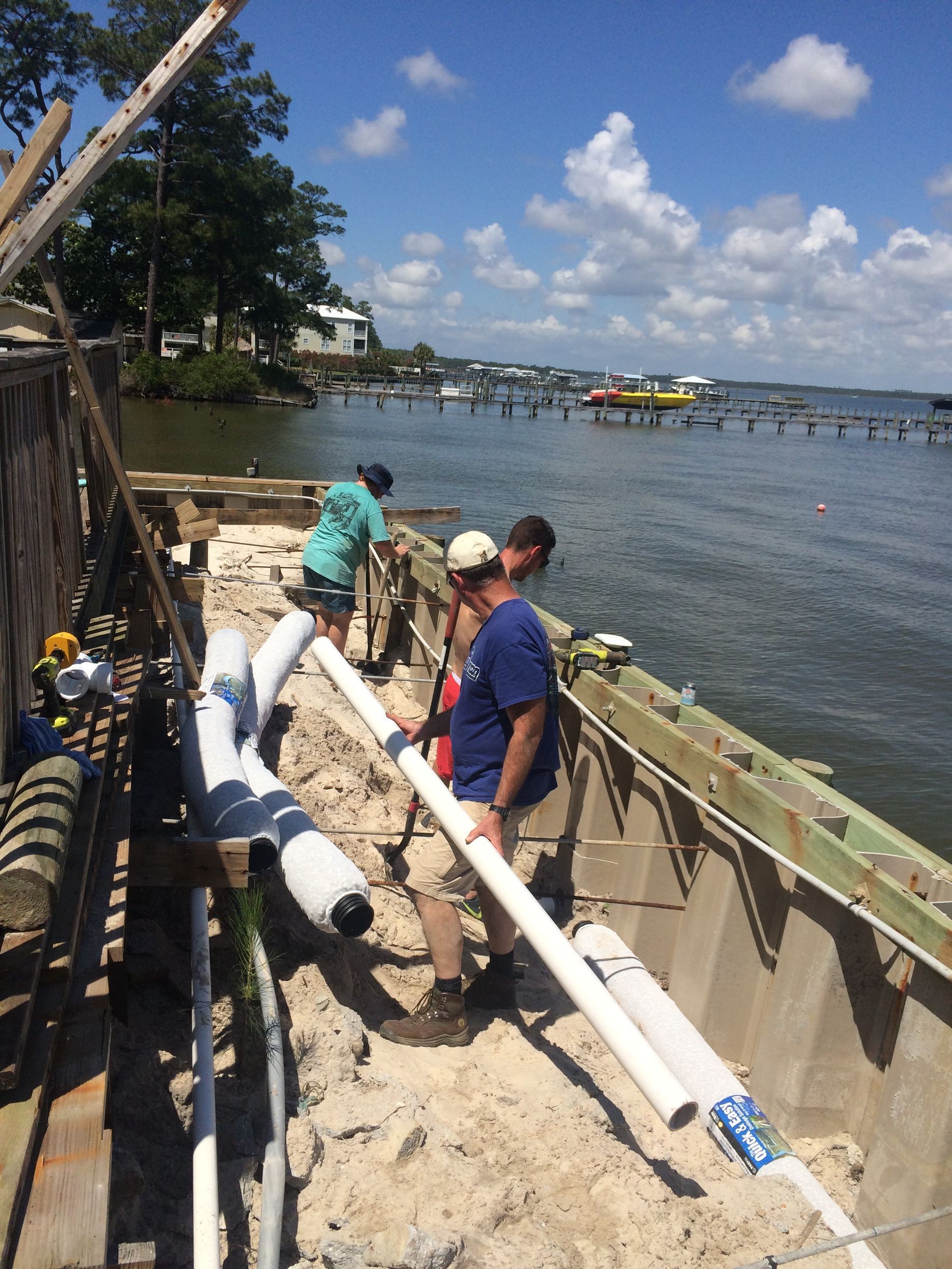 Three workers install white pipes along a concrete waterfront structure, near a body of water under a bright sky.