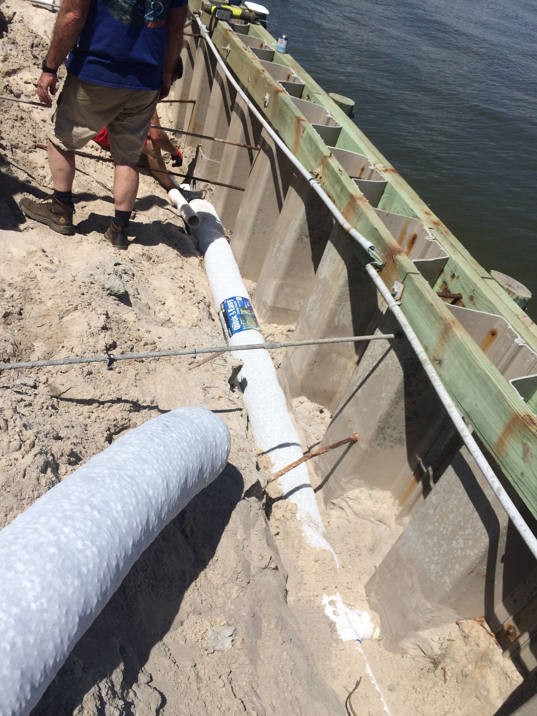 Construction workers installing a white wrapped pipe on a concrete structure near water.