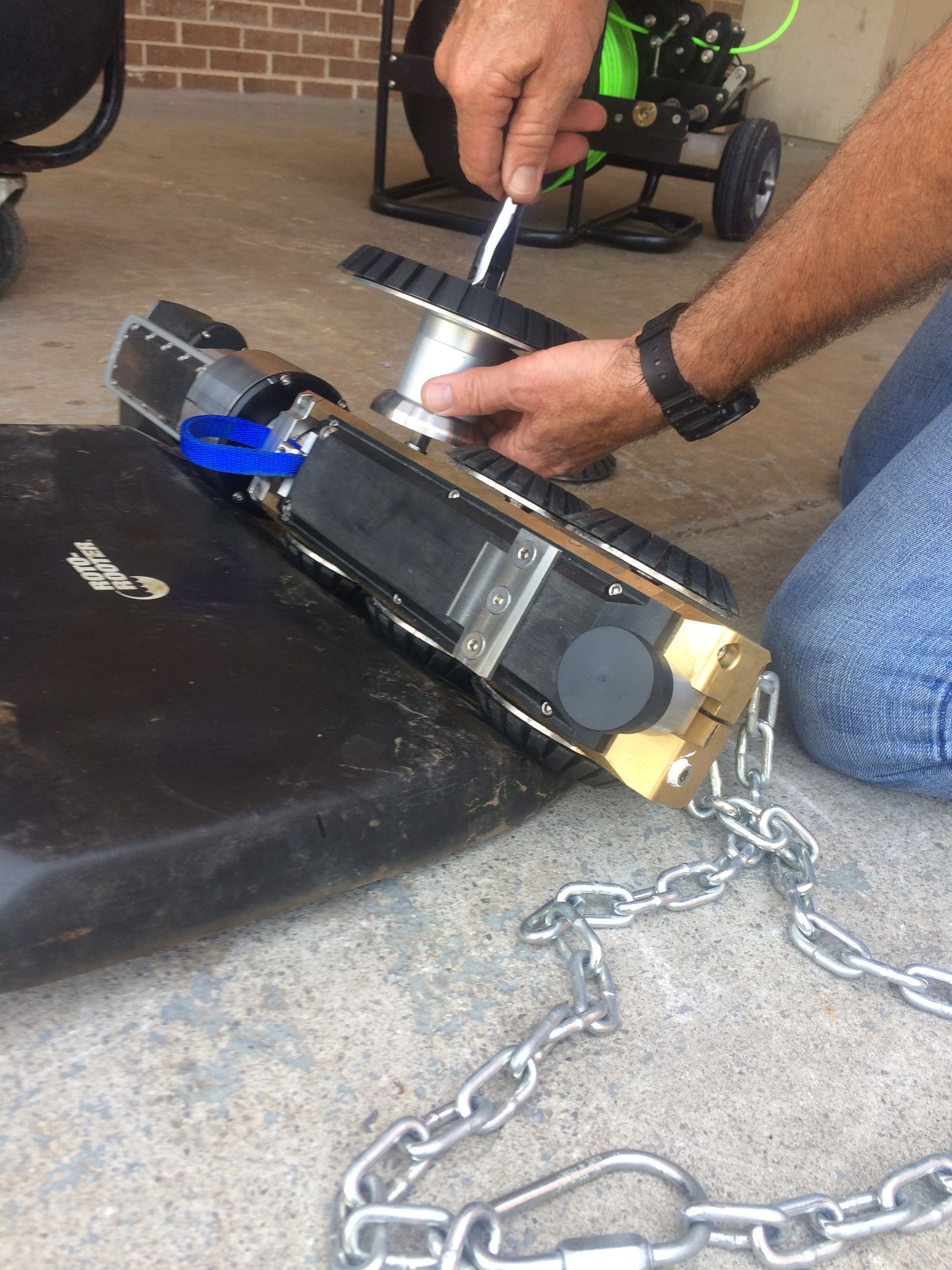 Person attaching a wire spool to a crawler robot with a chain, outdoors.