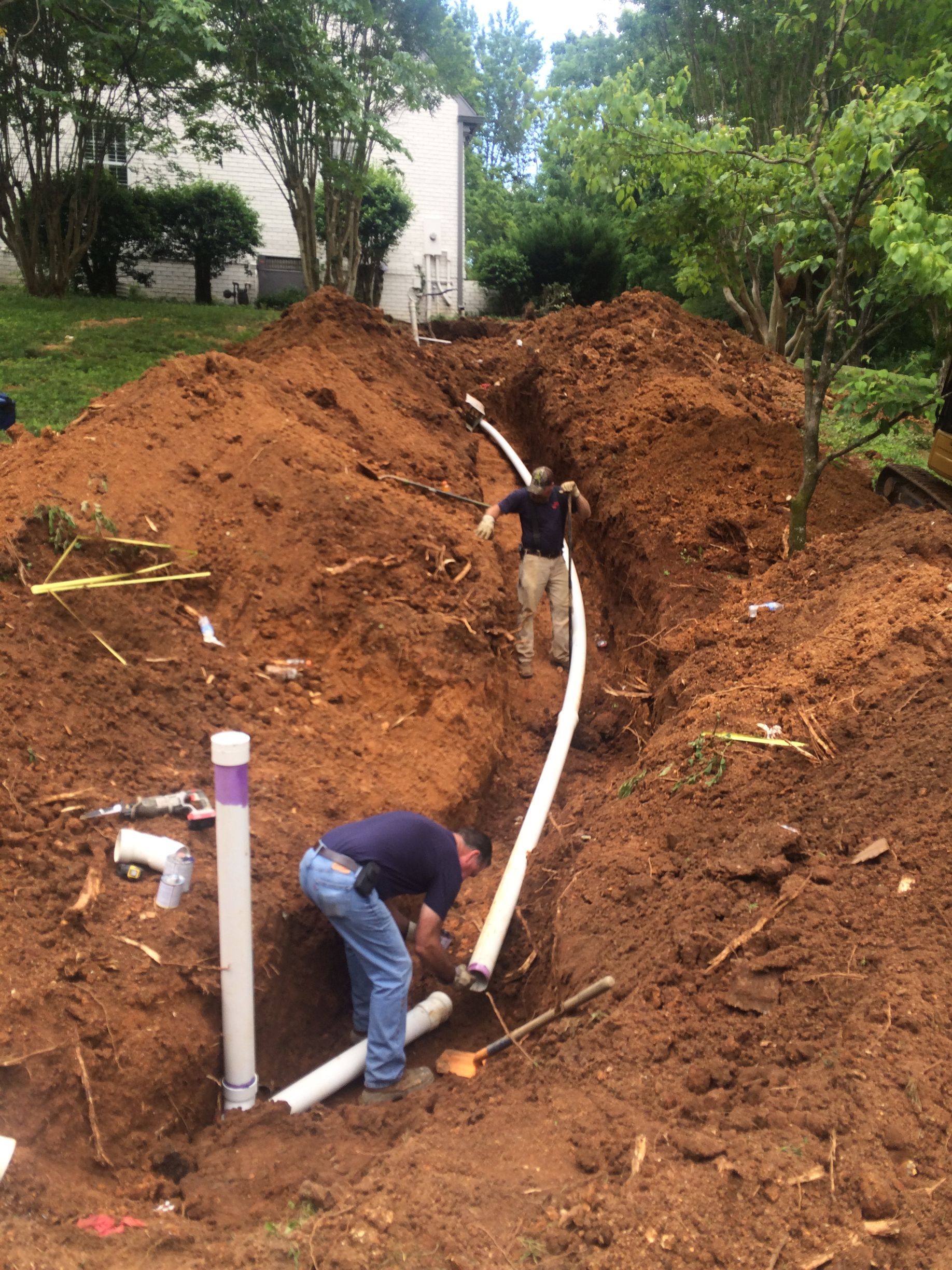 Two workers installing pipes in an outdoor trench, surrounded by dirt.