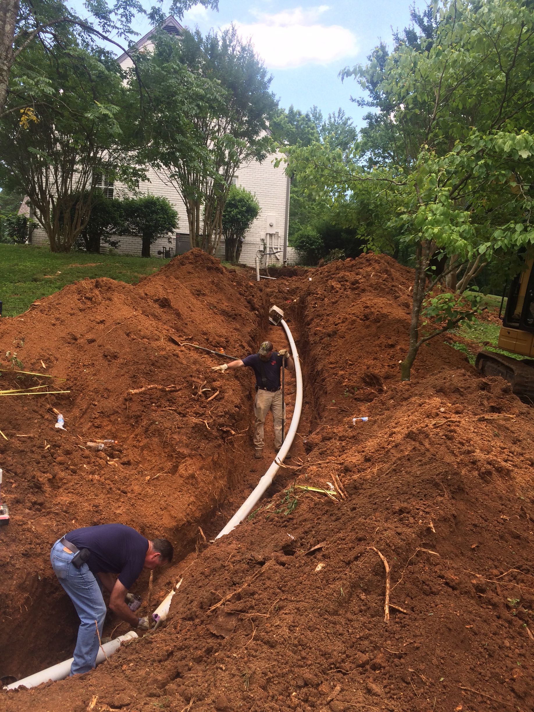Men installing white pipes in a trench on a hillside. Red dirt surrounds them, trees in the background.