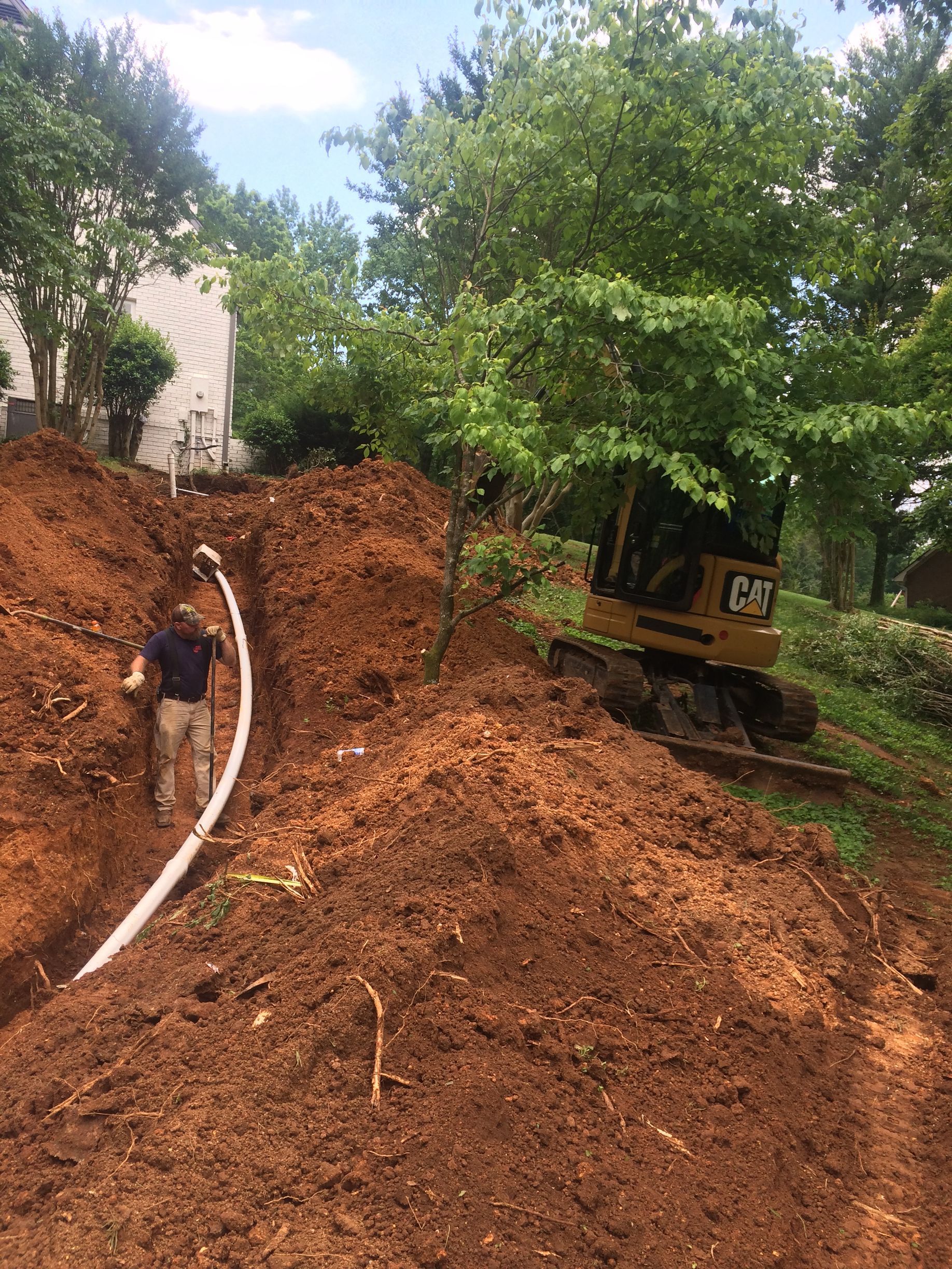 A person installing white pipe in a trench on a hillside with a CAT excavator.