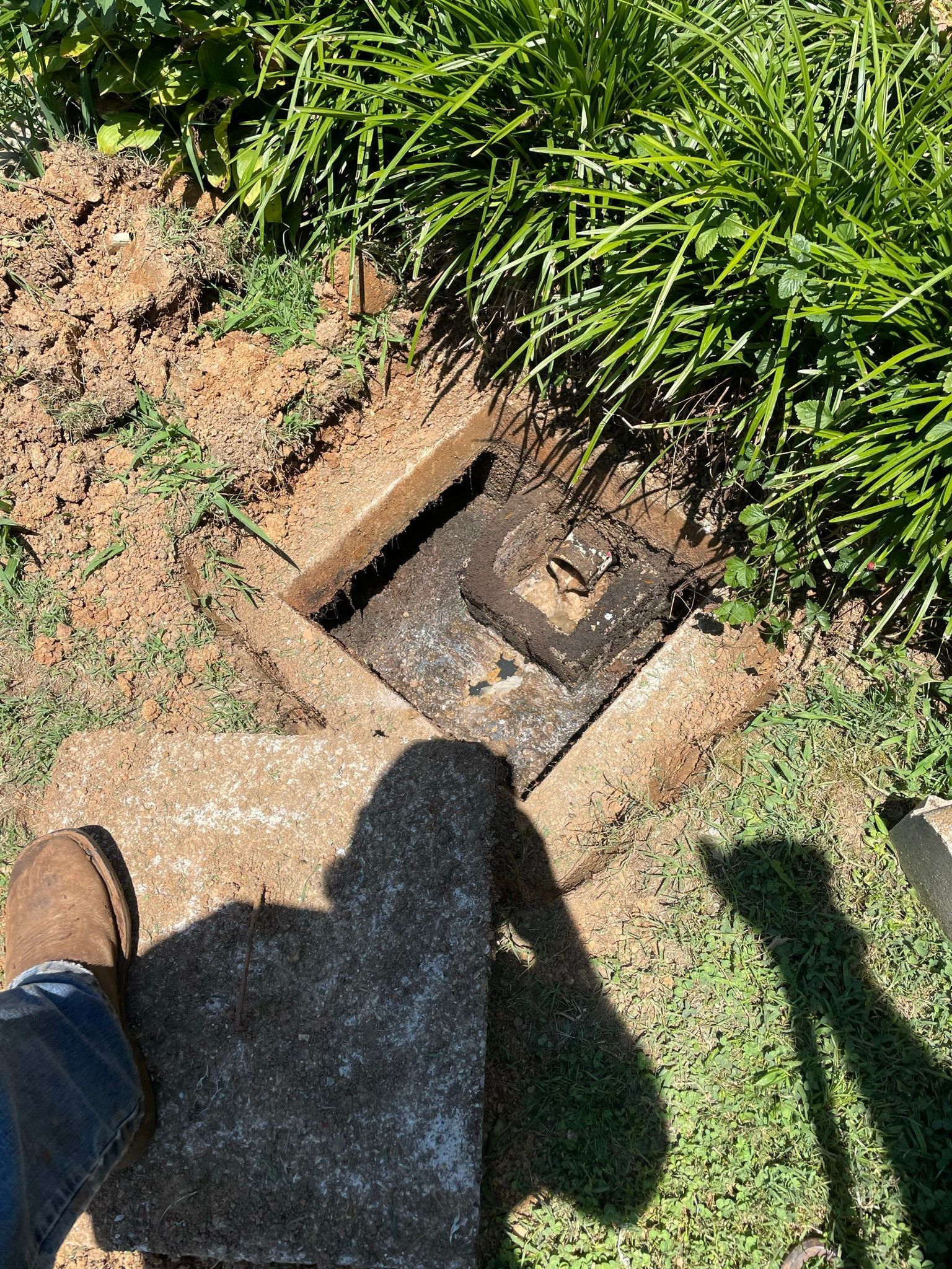 Open square concrete access point in dirt and grass. A person's shadow is cast on concrete.