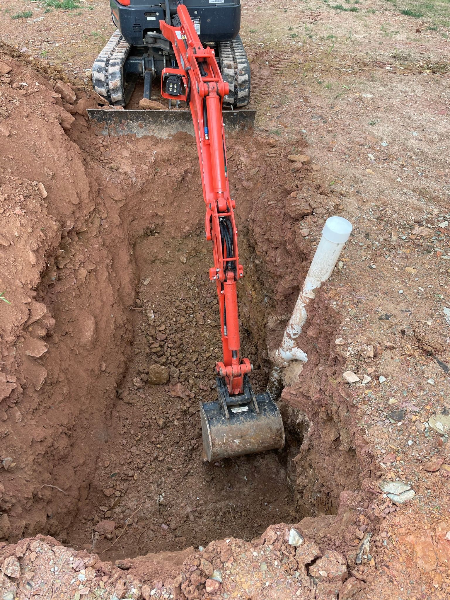 Mini excavator digging a trench near a white pipe, in a dirt setting.