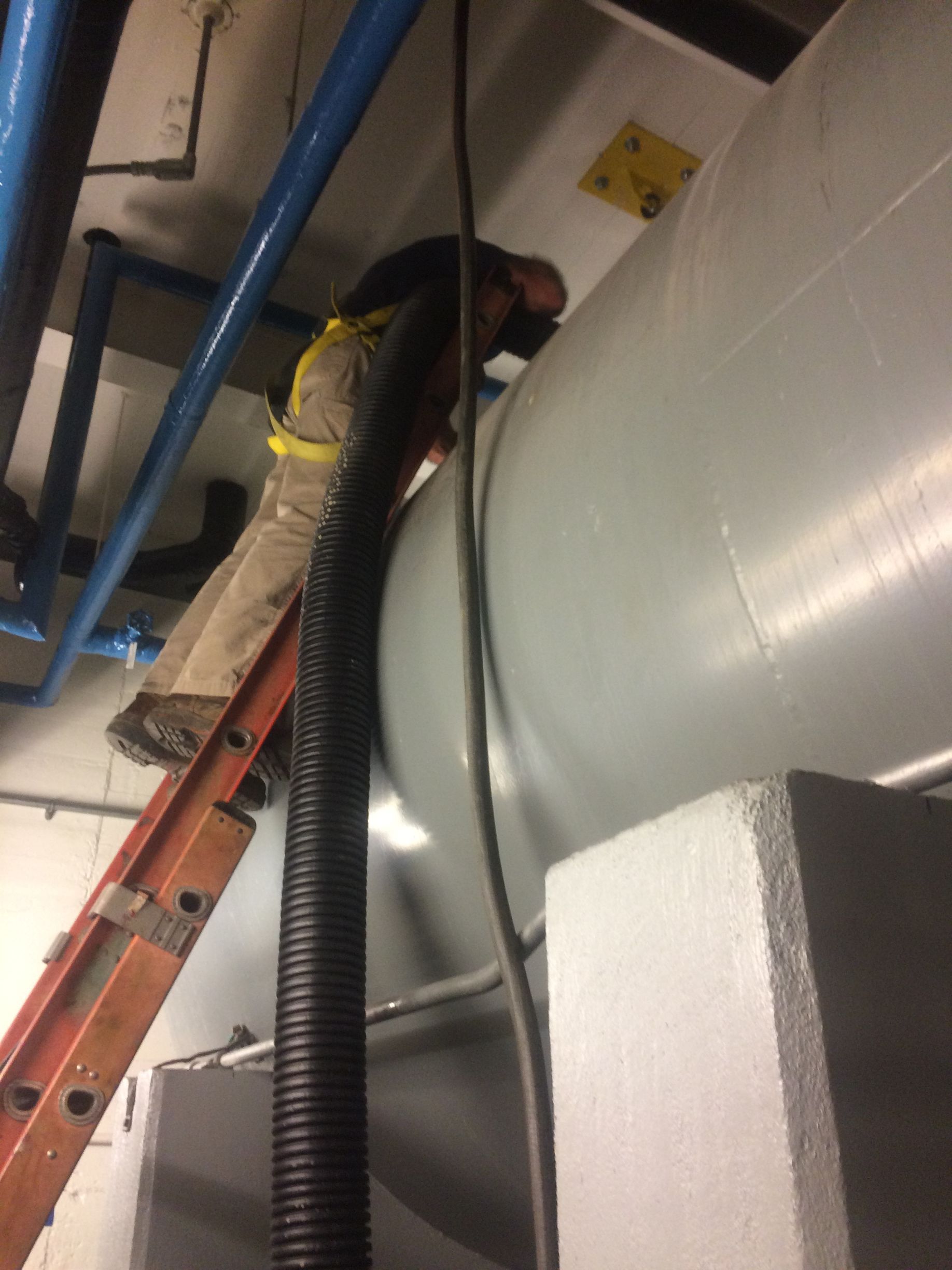 Person on a ladder working on a large, white cylindrical tank. Black hose runs down. Pipes and ceiling in background.
