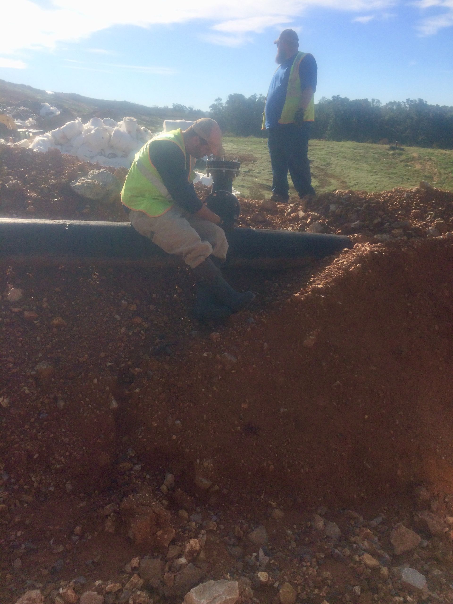 Two workers in vests near a trench, one working on a pipe, other observing in daylight.