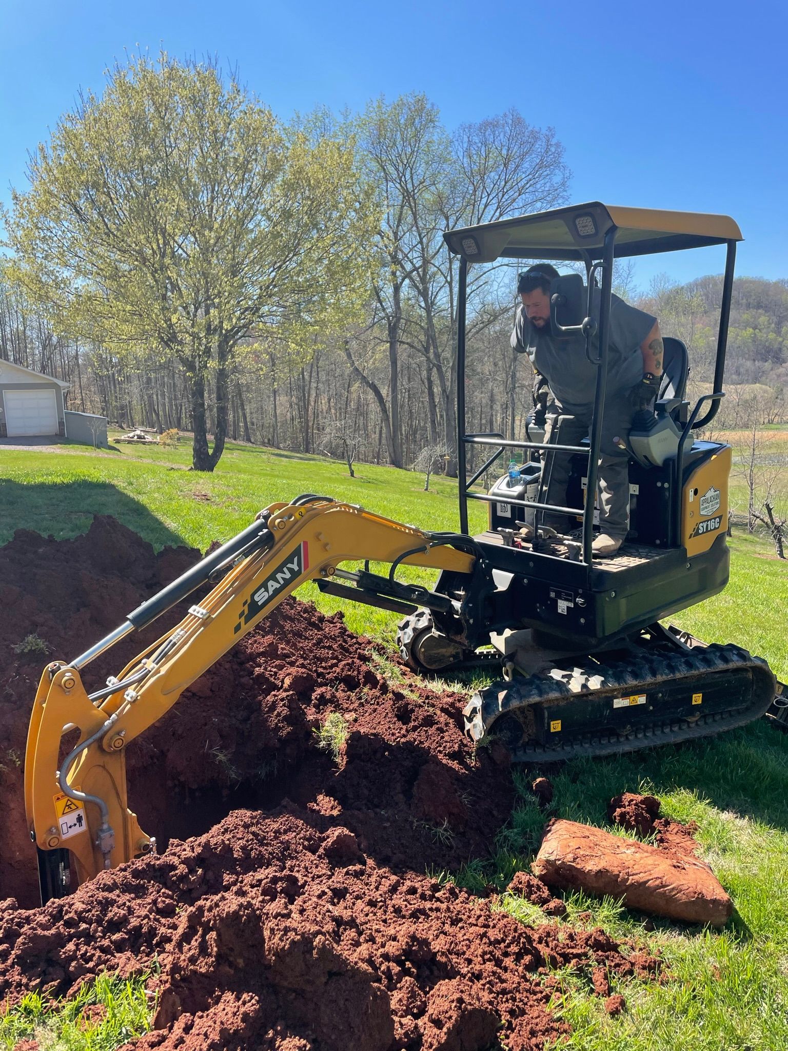 A person operating a yellow excavator digging a trench in a grassy area.