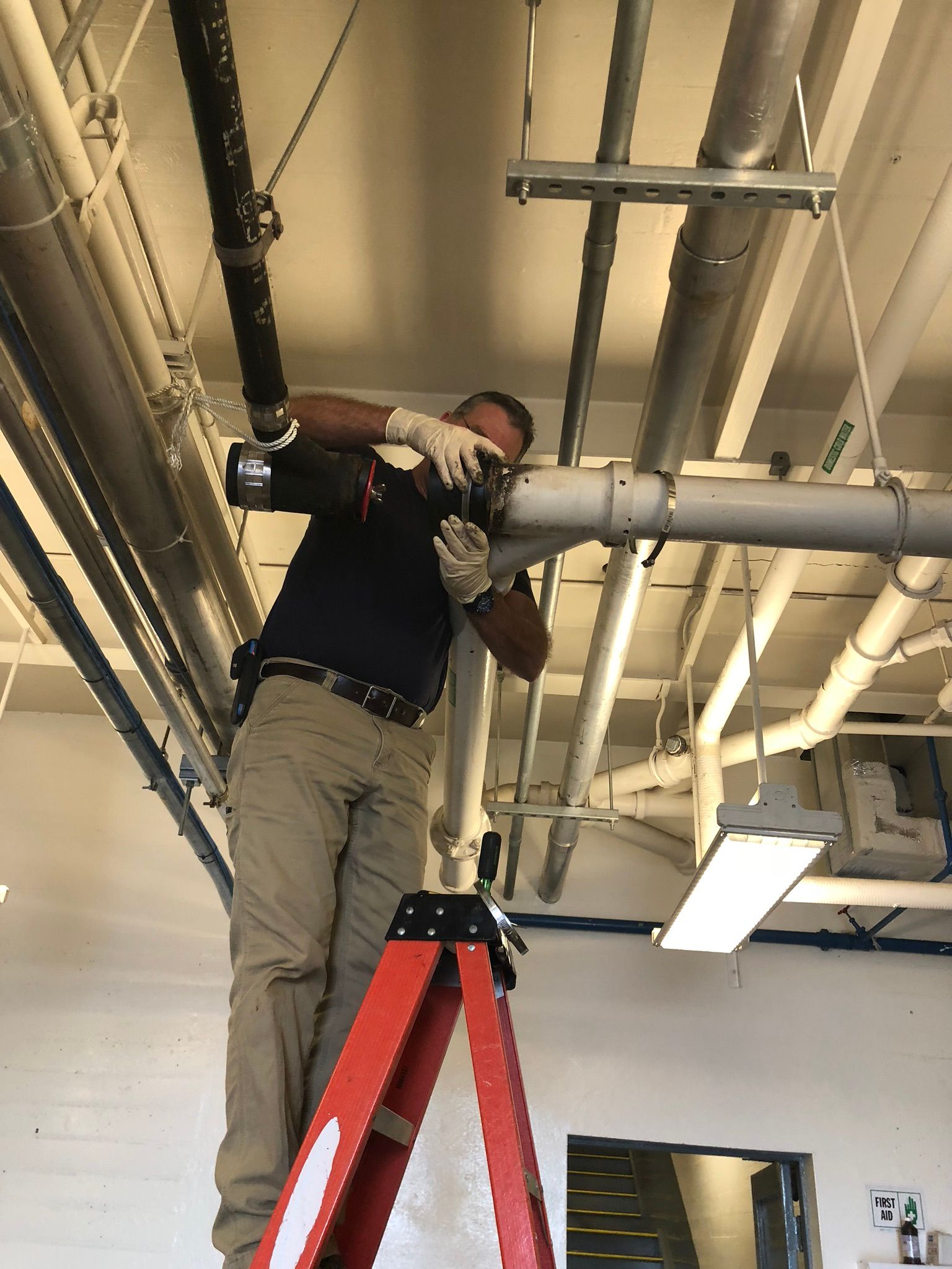 Man on a stepladder working on pipes in a building, wearing gloves.