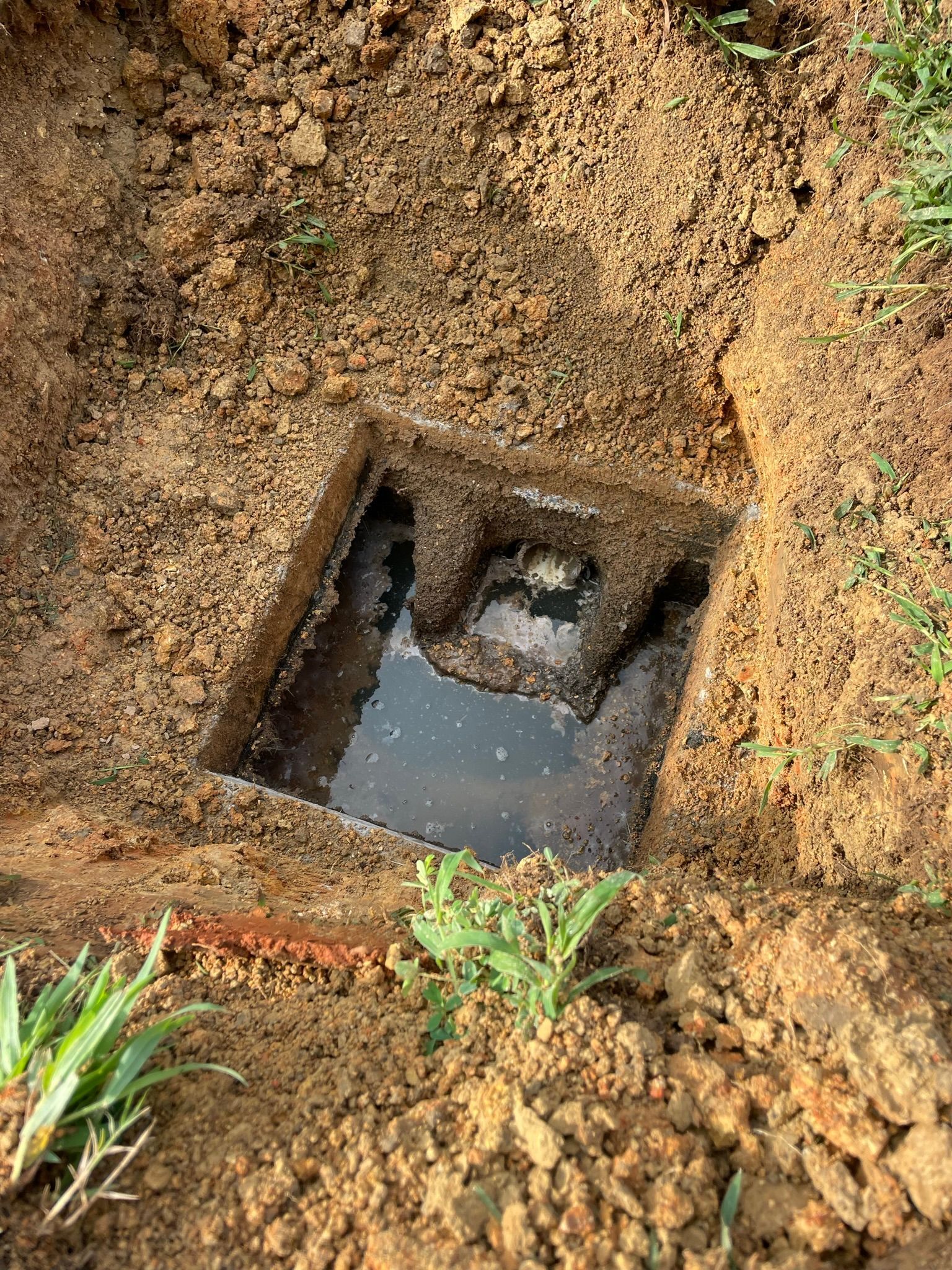 Open square hole in the ground with dark water pooling at the bottom, surrounded by soil and grass.