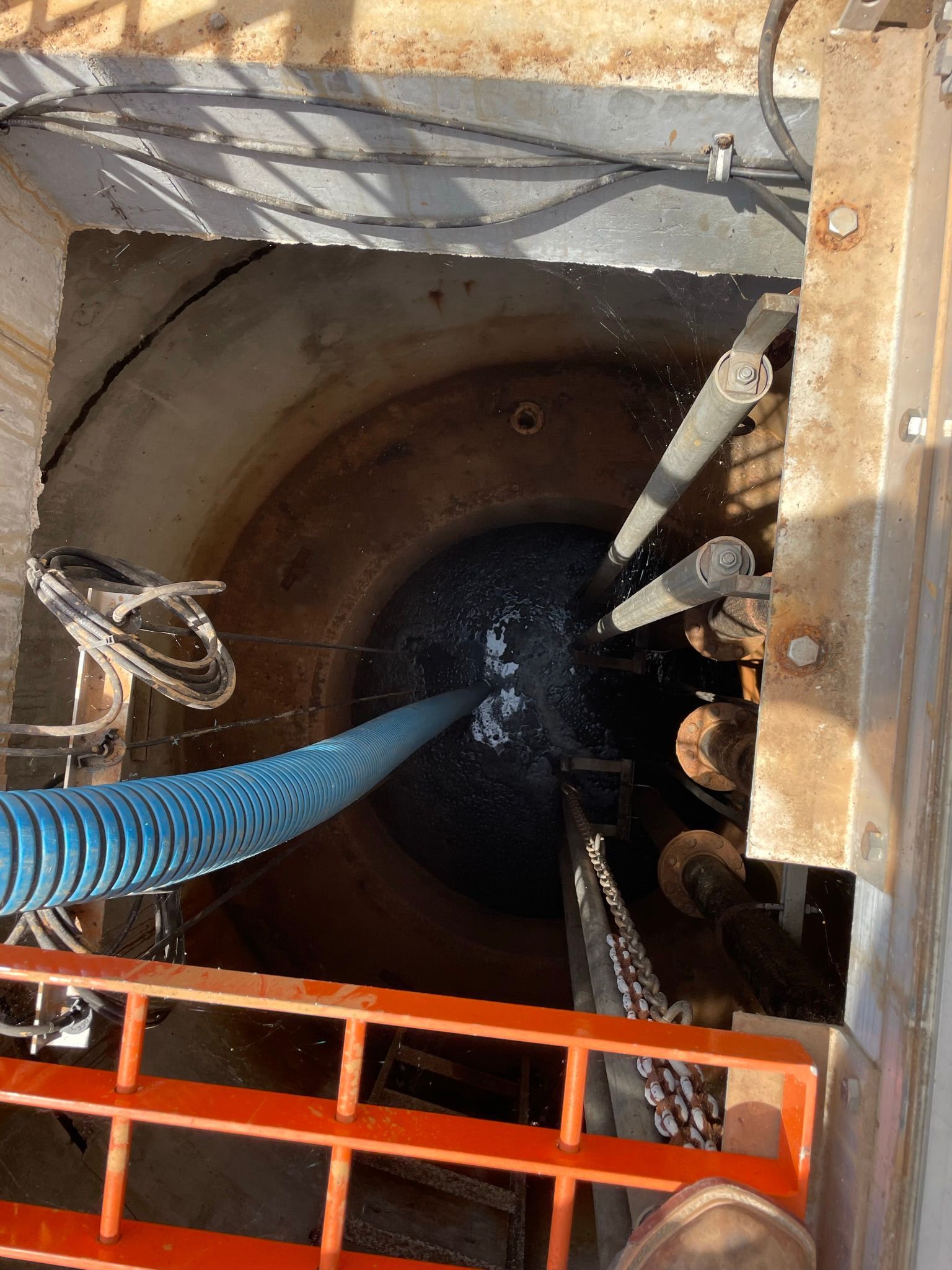 Looking down a deep well, blue hose, pipes, and water are visible. Orange railing surrounds the opening.