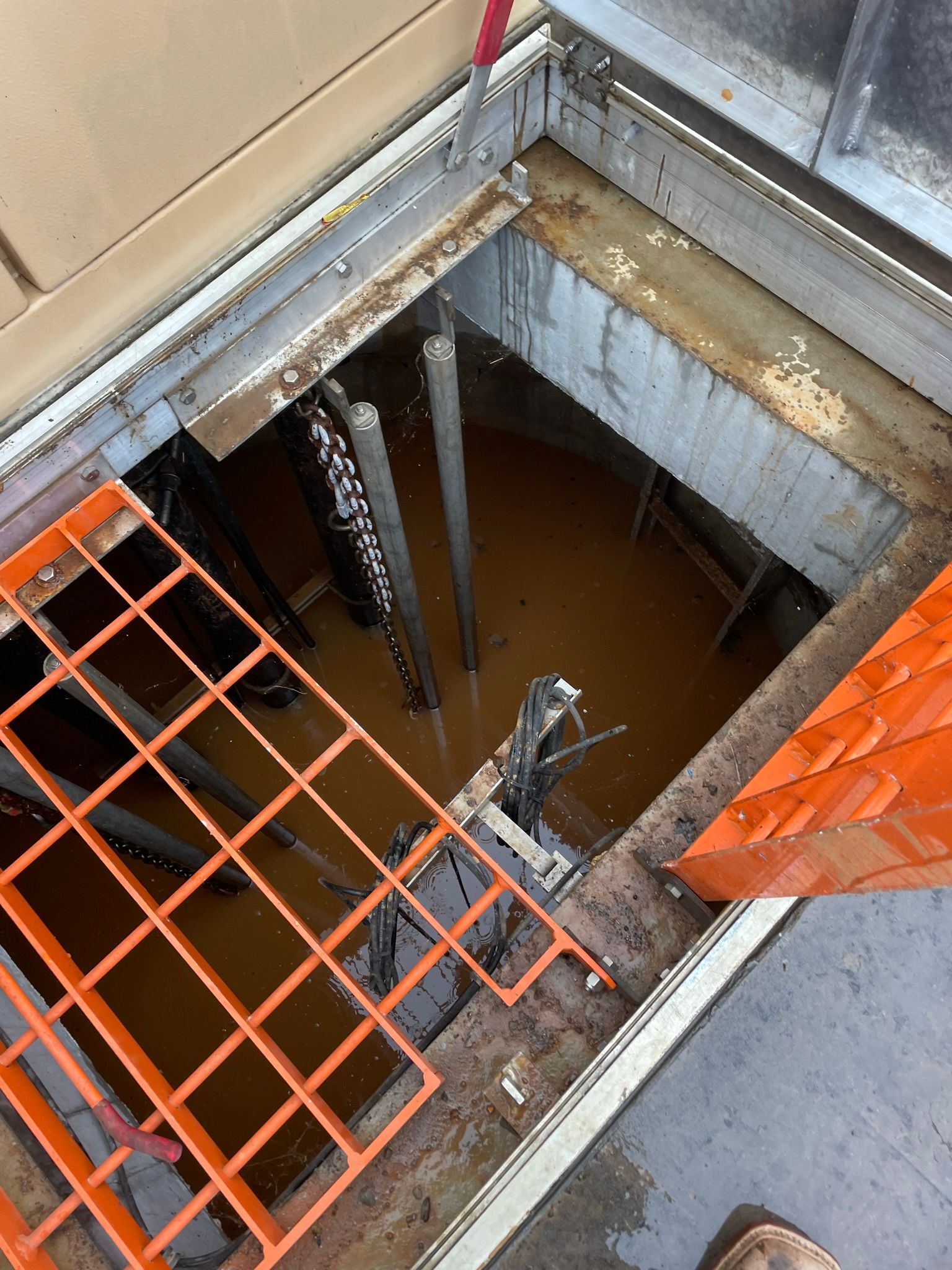Orange grates and a metal frame surround a flooded pit. Wires and pipes hang down into the murky brown water.