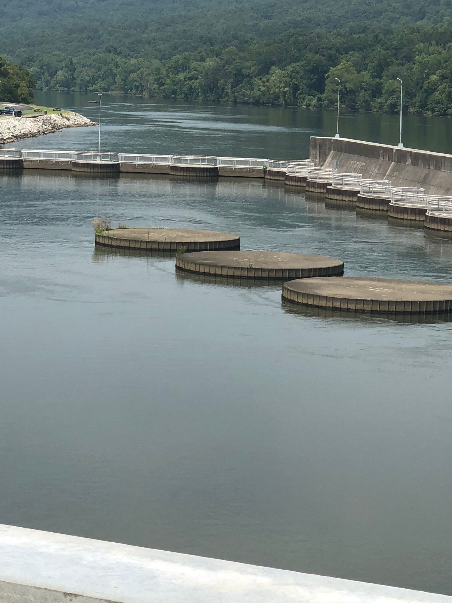River with stepping stones, dam, and forested hills in the background.