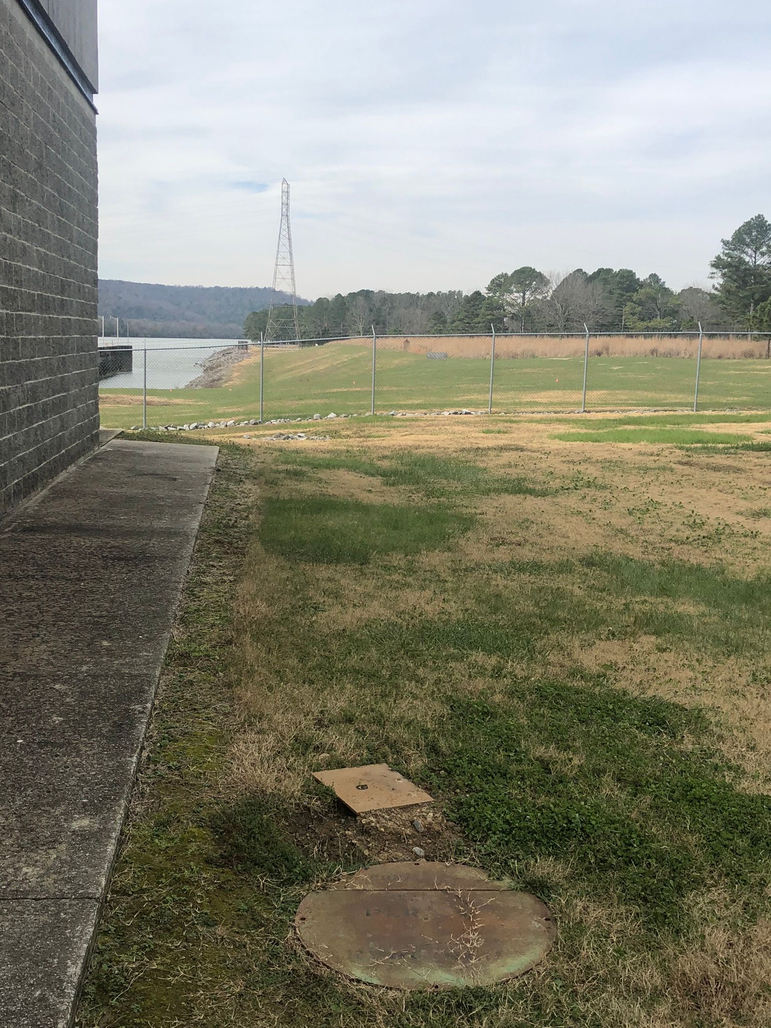 Grassy field with a lake and distant tower visible beyond a brick wall and sidewalk. Overcast sky.