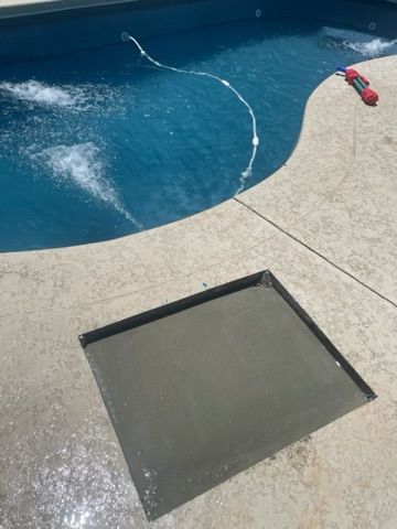 Concrete-filled square cutout in a concrete patio next to a swimming pool with jets.