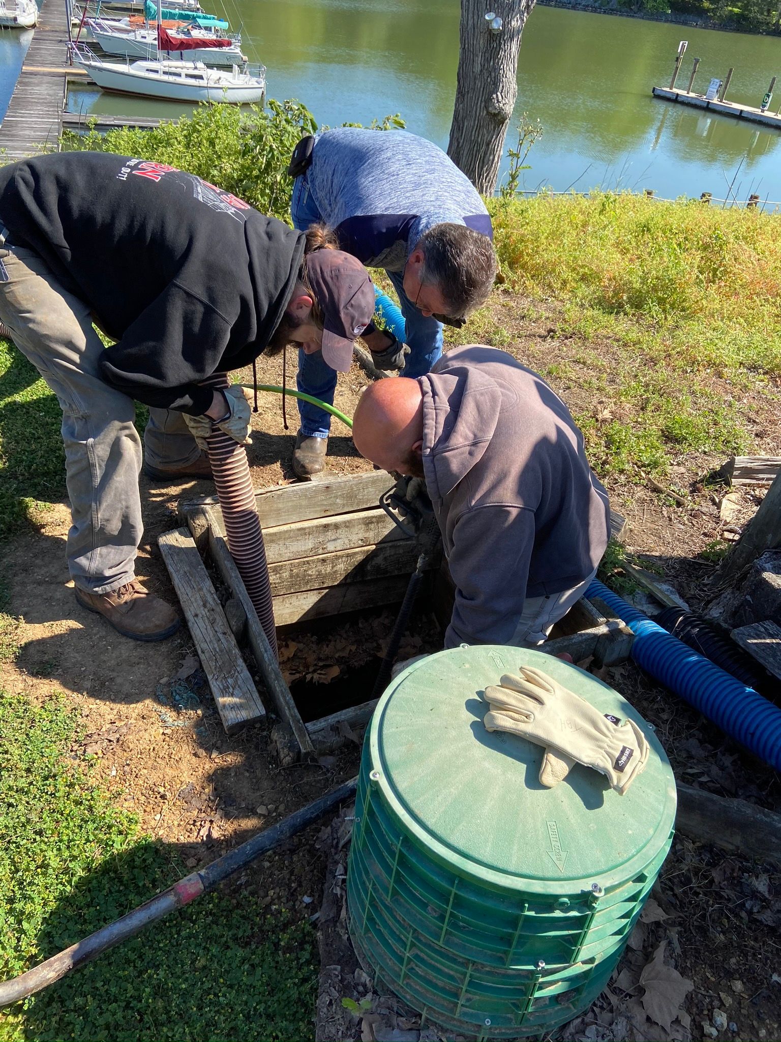 Three people working on an open access point near a body of water and boats; one holding a tool, another looking down.