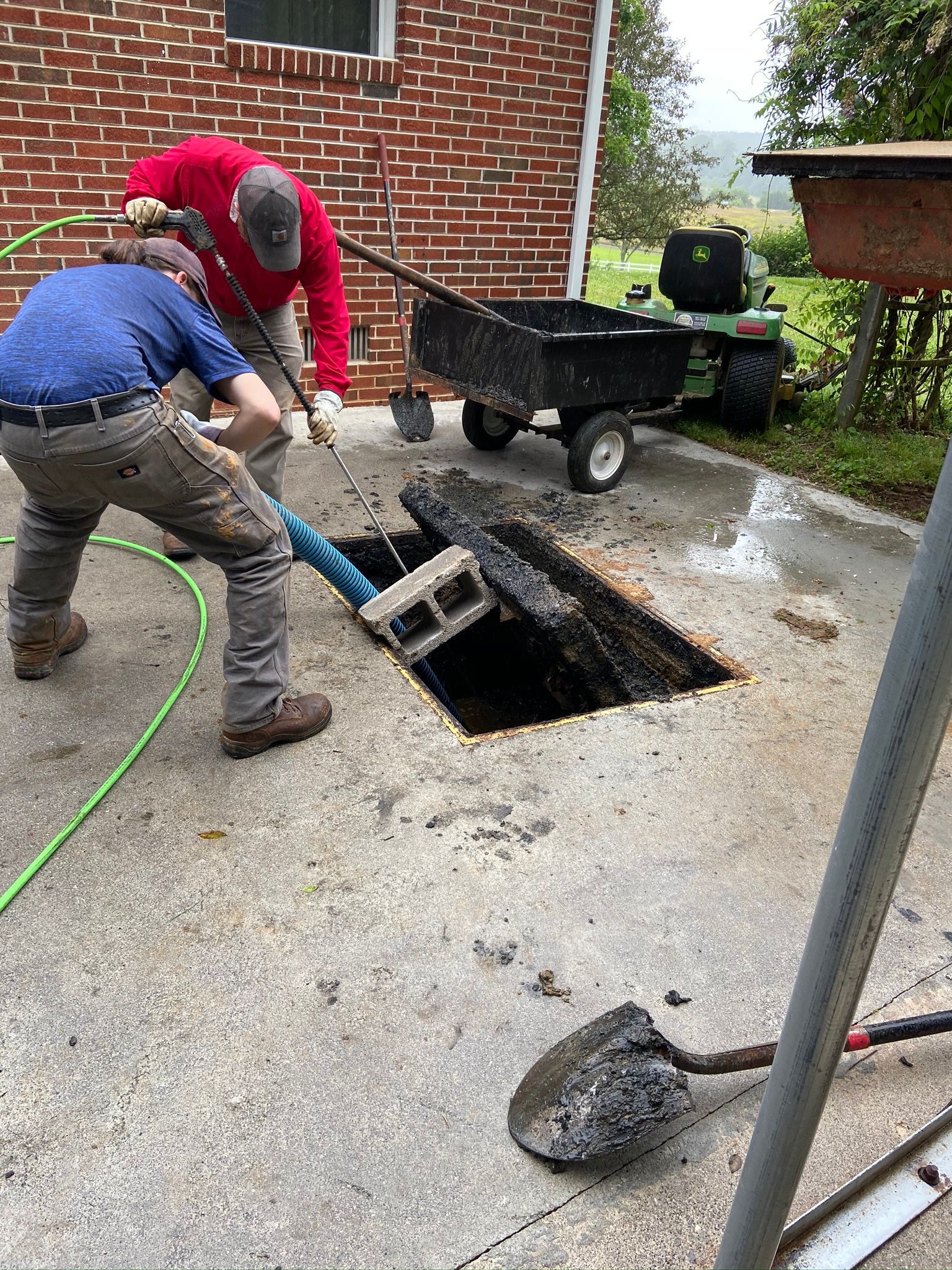 Two people lifting a concrete slab from a hole in a concrete surface. A cart and shovel are nearby.