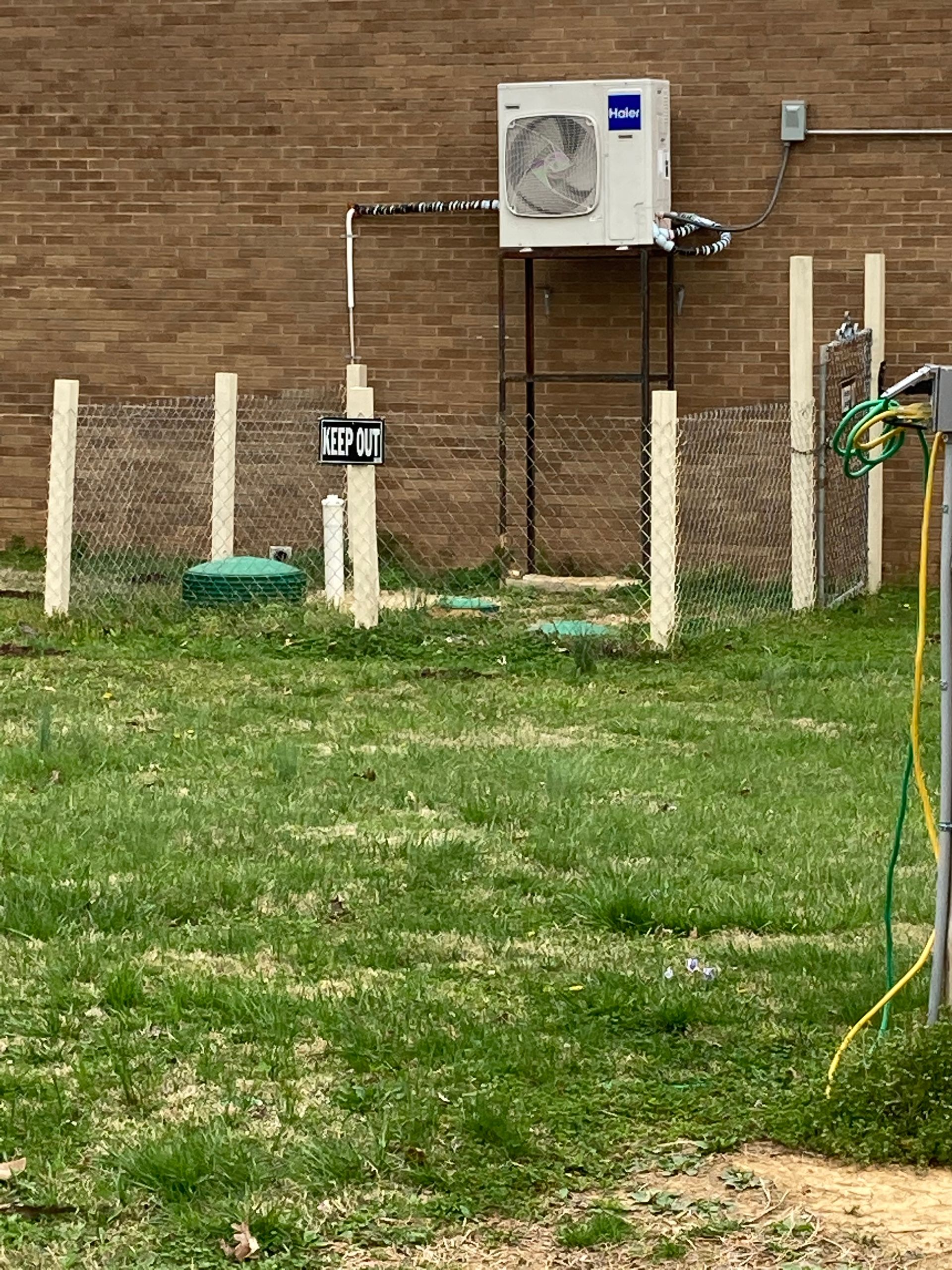 Air conditioning unit mounted on a brick wall, enclosed by a chain-link fence, grass, and a 