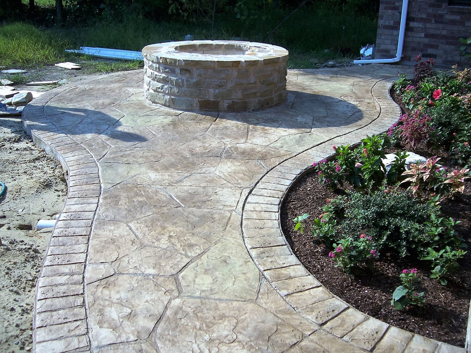 Stone fire pit on a stamped concrete patio with a brick border, next to a flower bed.