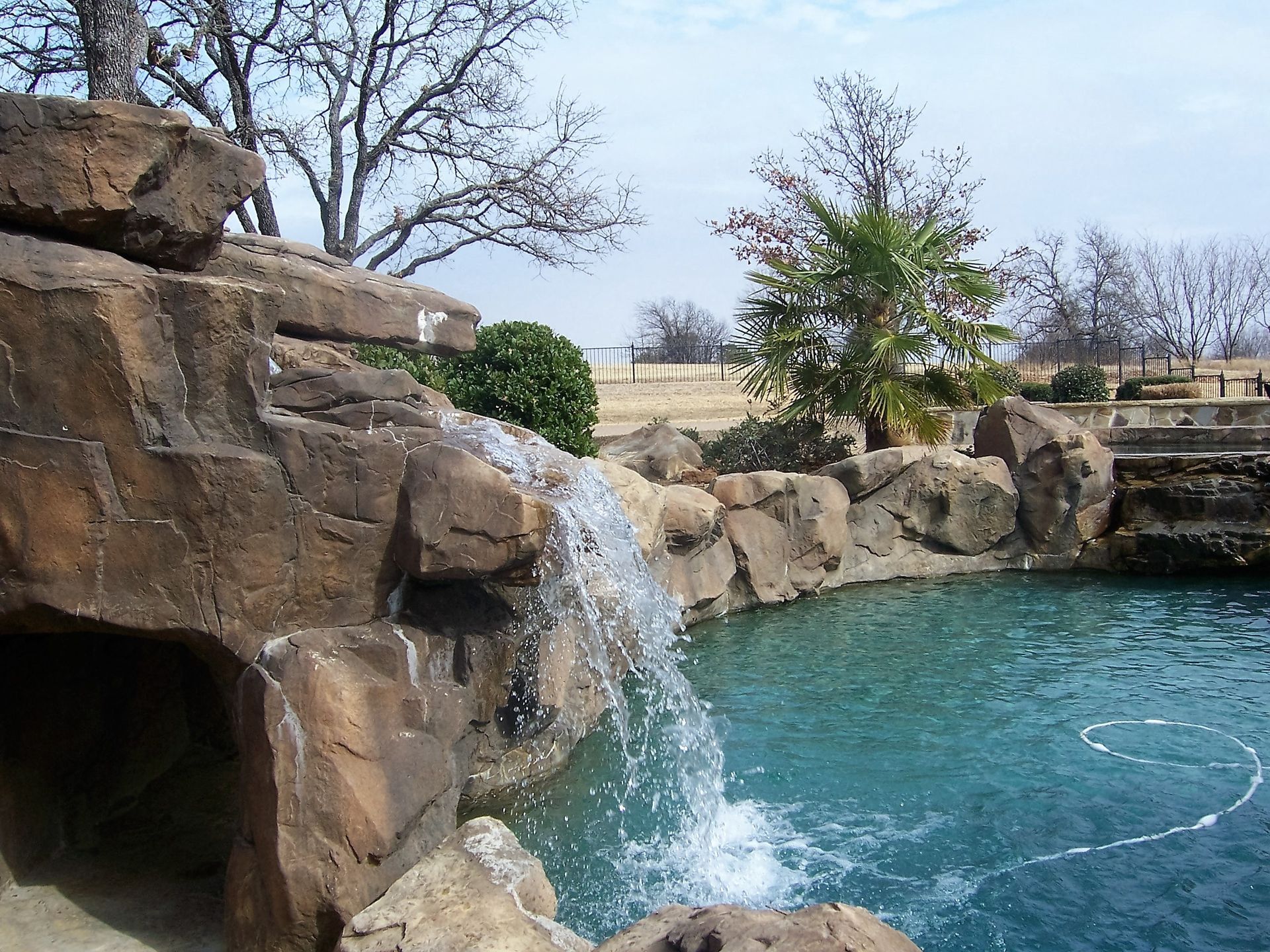 Waterfall cascading into a turquoise pool surrounded by rocks. Trees and a fence are in the background.