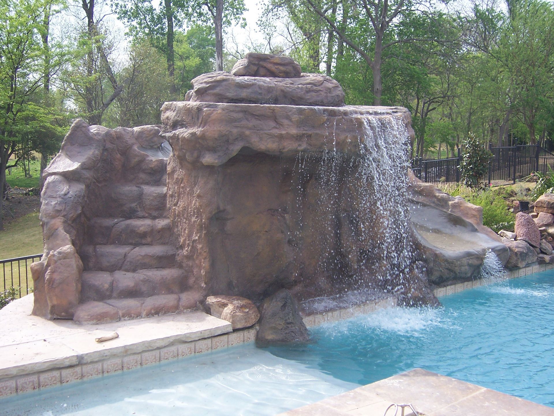 Pool with a rock waterfall, steps, and a small slide; blue water and trees in the background.