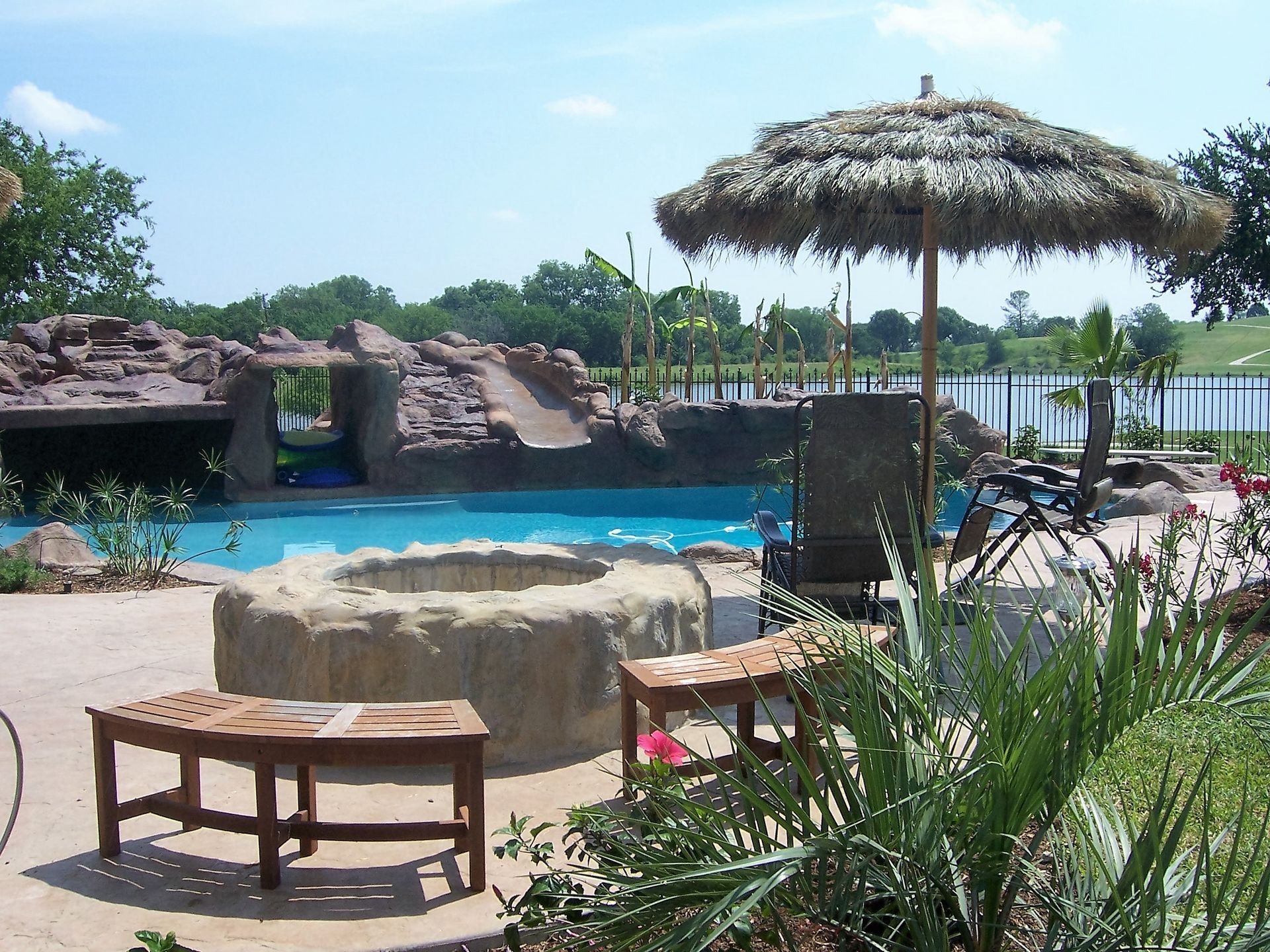 Poolside scene with tiki umbrella, lounge chairs, rock features, and lush greenery.