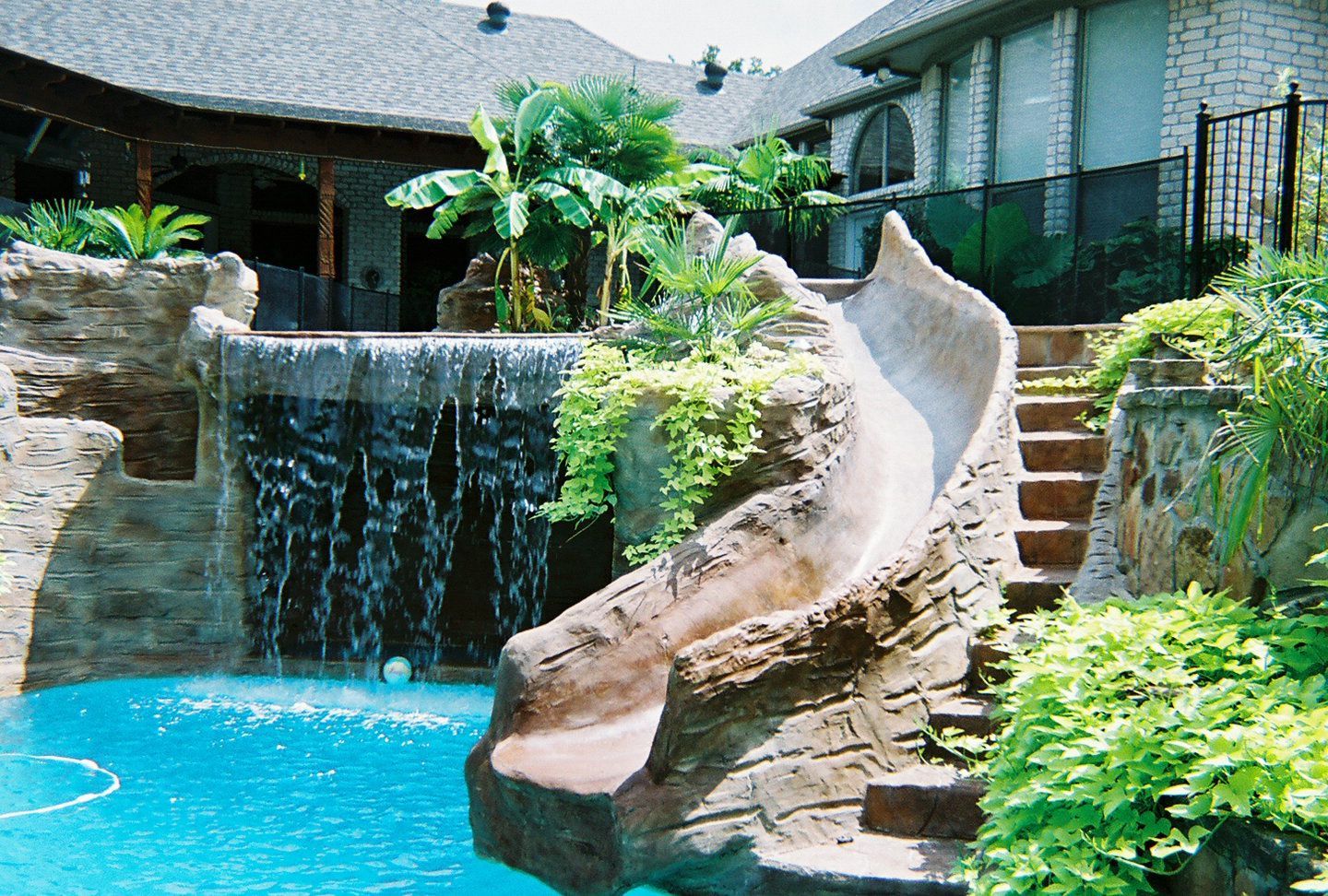 Pool with waterfall, rock slide, and lush landscaping in backyard of a home.