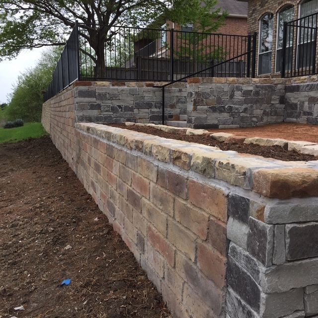 Stone retaining wall with black metal fence and steps, separating a yard from a house and garden.