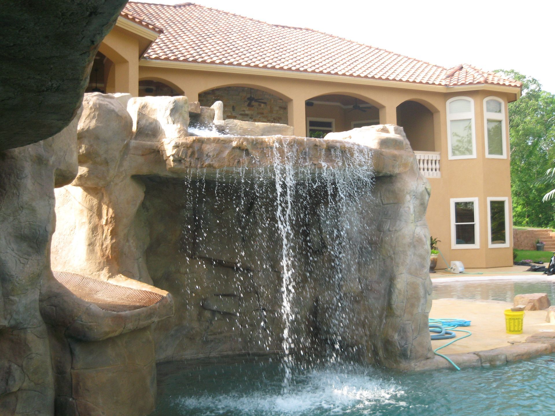 Waterfall cascading into a pool in front of a two-story beige house with a red-tile roof.