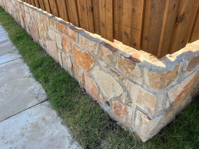 Stone wall with brown wooden fence behind it, beside a sidewalk and grass.